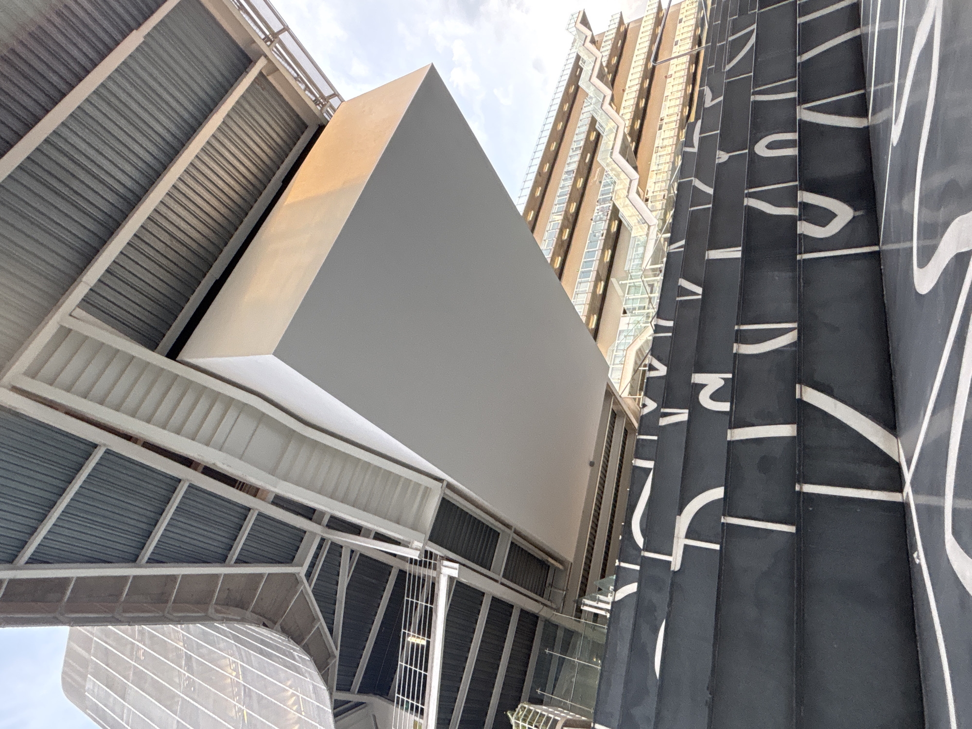 The image presents an upward perspective of architectural elements in Mexico City, Mexico. Dominating the foreground is a large, rectangular white structure, positioned adjacent to a building with dark, vertically aligned panels adorned with white, curvilinear designs resembling stylized lettering. To the left of the white structure, a metal framework with a corrugated underside suggests a walkway or overhead shelter. In the background, a taller building is visible, featuring a grid-like facade with glass and concrete or stone elements. The sky is partially visible in the upper background, appearing light and overcast.