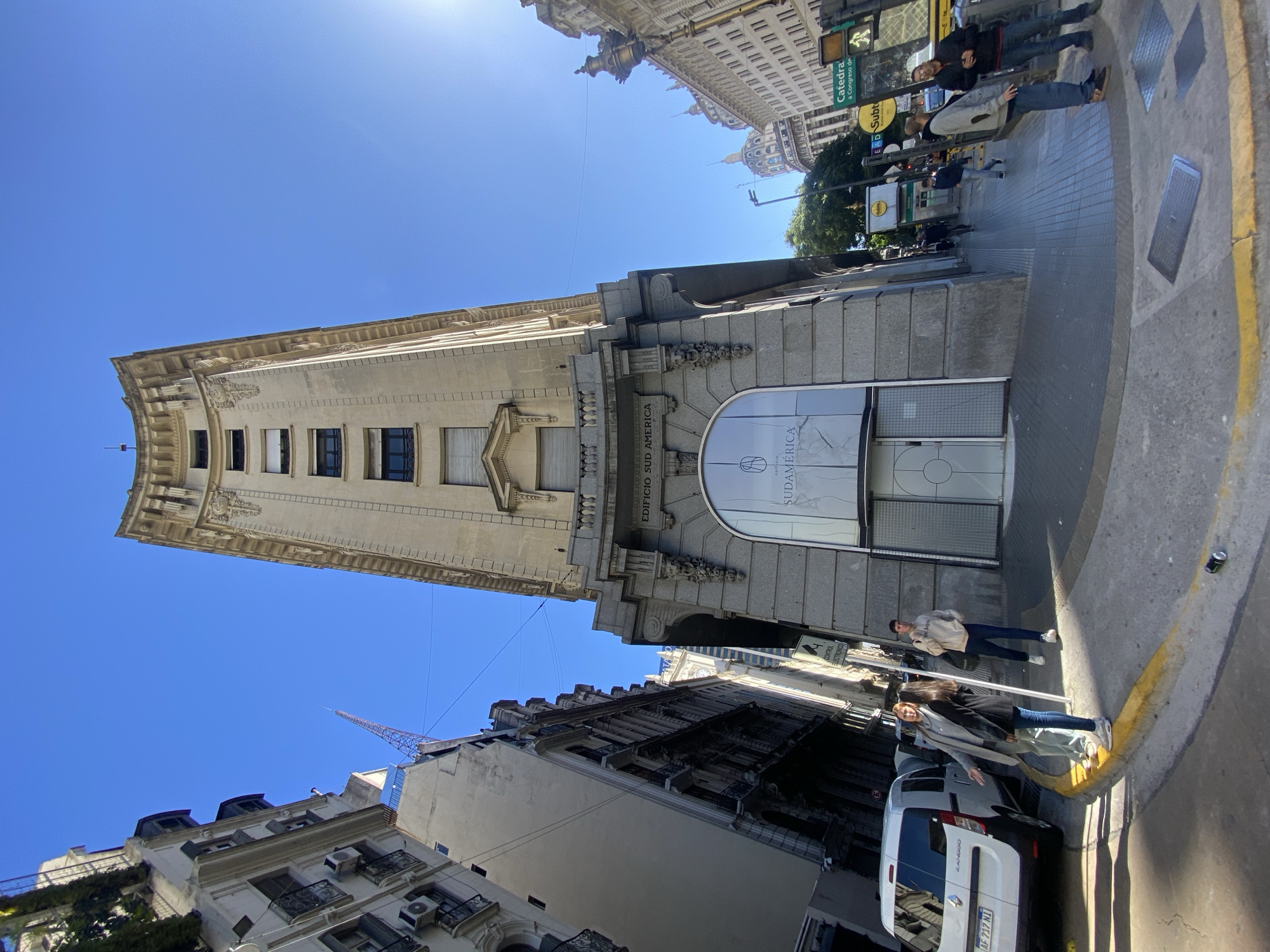 A medium-angle shot taken from a low angle depicts the Edificio Sud América in Buenos Aires, Argentina. The building's facade is made of light-colored stone, featuring a central, slender tower rising above a lower, wider section. The entrance features a large arched window with "SUDAMERICA" displayed prominently. The building's style incorporates classical architectural details.
The foreground shows a section of a sidewalk with a yellow curb and people walking, with a white van parked at the curb, partially obscured by the passers-by.
In the background, buildings line the street, and traffic signs are visible. The sky is clear and blue, with a bright sun. The angle of the shot emphasizes the height of the building.