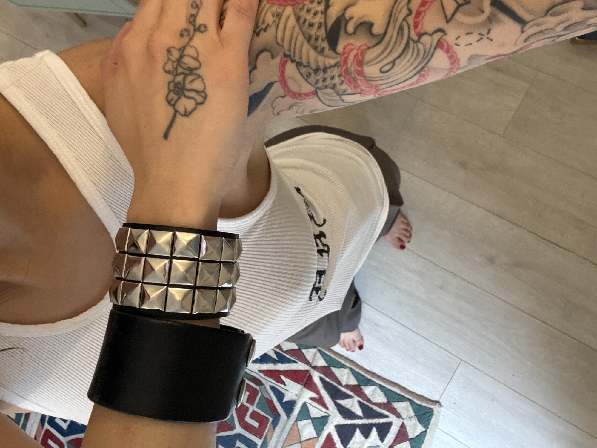 An overhead shot depicts a person wearing a white tank top and gray bottoms. They have tattoos on their arms and a tattoo on their hand. The hand with the flower tattoo is close to the neck. The person is wearing a wide black cuff bracelet and a silver studded cuff bracelet on their wrist. Parts of the person's feet are visible. A patterned rug is on the floor. The background is a light-colored hardwood floor. This image does not contain sufficient information to identify a location in London.