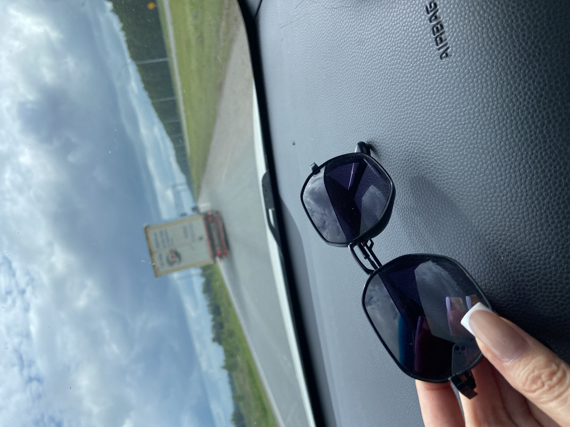 An image taken from inside a vehicle shows a pair of dark-rimmed sunglasses resting on the dashboard. The sunglasses are positioned in the foreground and reflect the cloudy sky and passing landscape. In the background, a white truck with a red trailer is visible on a highway. The truck is moving away from the camera. The road is flanked by green trees and a grassy shoulder. The sky is overcast with a mix of light and dark clouds. The word "AIRBAG" is embossed on the dashboard to the right of the sunglasses. The overall impression is of a journey on a road under a moody sky. The location is identified as the city of Mirny, Russia.