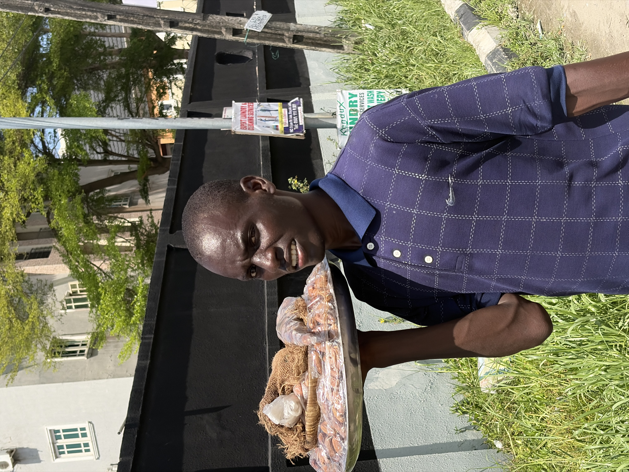 In Igboefon, Nigeria, a middle-aged African man is captured in a medium shot, holding a large oval tray laden with items, including packaged goods, possibly food items. He wears a dark, short-sleeved, button-down shirt with a subtle grid pattern. His expression is slightly strained, as if the tray is heavy.
Behind him, a black fence stretches along the property line, beyond which is a partial view of a multi-story building with windows. To the left, there's a utility pole with multiple signs affixed to it, and grass covers the ground to the right. The lighting indicates it is daytime, with sunlight casting shadows. The overall setting appears to be an urban or suburban area.