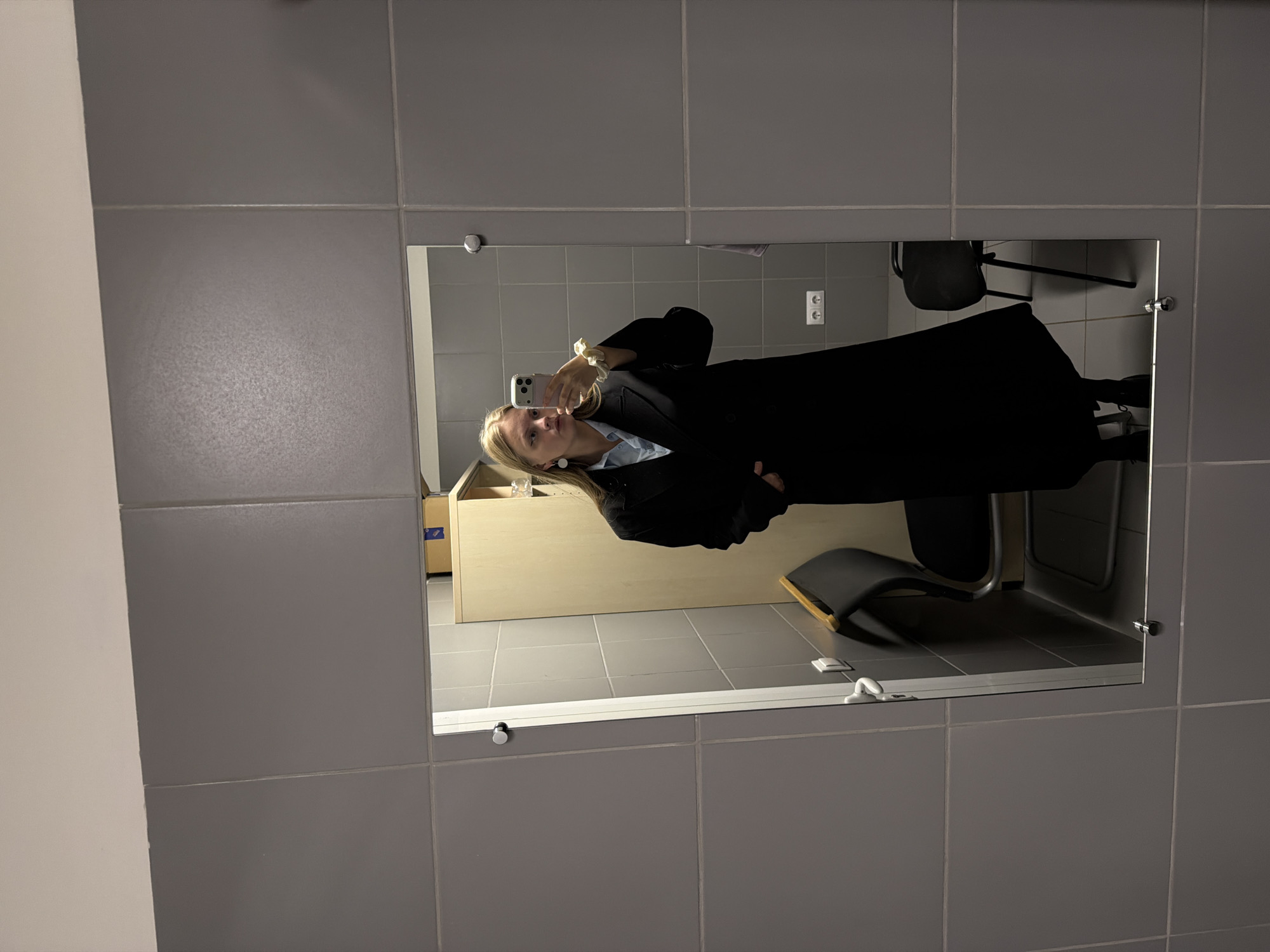 The image is a reflection in a square mirror mounted on a tiled wall with gray tiles. The reflection shows a young woman taking a photo of herself with her smartphone. She is wearing a black blazer and a light blue shirt. She has long blonde hair. Behind her, a light-colored wooden cabinet or drawer unit is visible. A black office chair with a gray seat is also visible. The scene appears to be indoors, potentially in an office or public space. The presence of an electrical outlet on the wall in the mirror suggests the location is in Russia. Specific to the city of Izhevsk, Russia, would require additional information beyond the visual content of the image, as no landmarks or identifiable features are present to conclusively determine the location.
