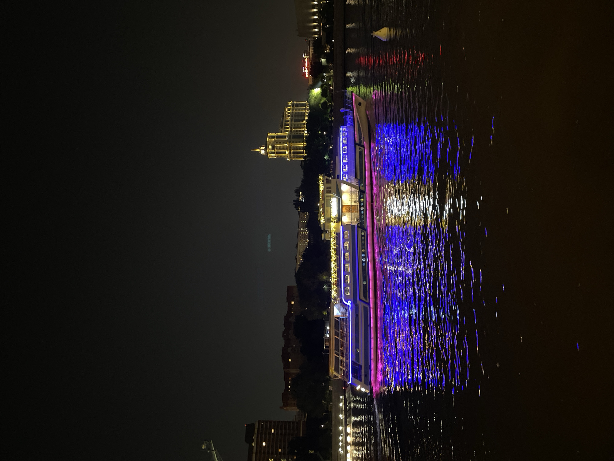 Captured at night, the image displays a vividly illuminated boat navigating the Moscow River. The boat features blue and pink lighting, which reflects brightly on the water's surface. In the backdrop, the cityscape of Moscow is visible, with buildings illuminated by warm lights. The overall setting presents a vibrant urban environment at night.