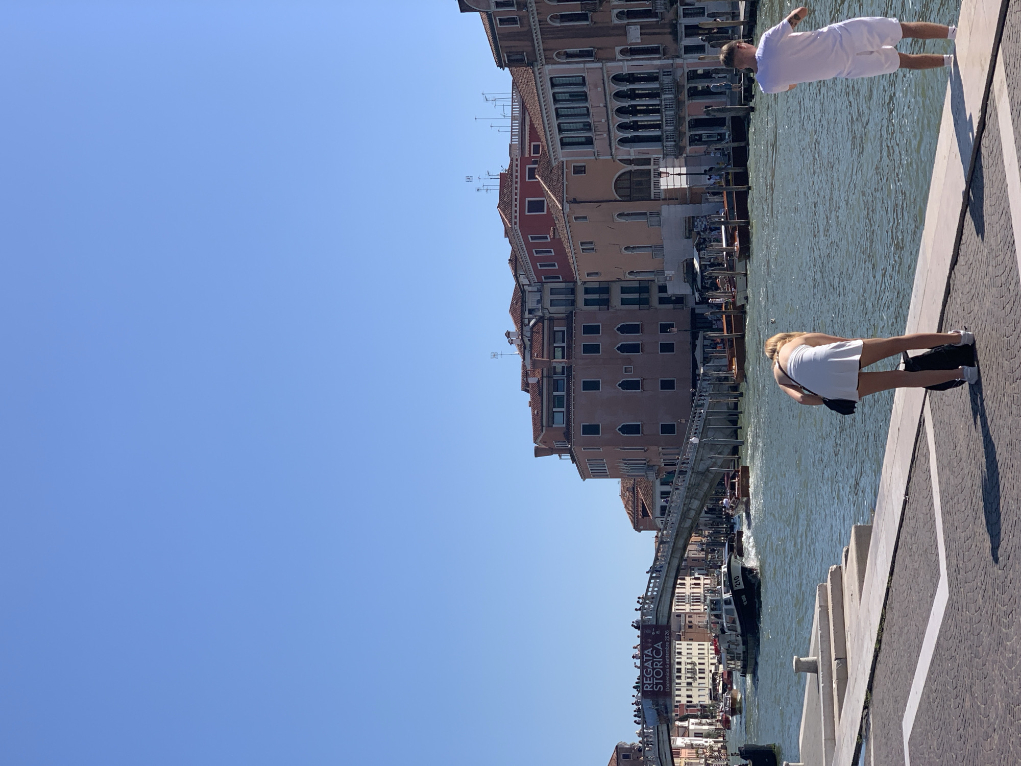 The image depicts a sunny day scene in Venice, Italy, likely on the Grand Canal, judging by the architecture and the presence of vaporettos (water buses) and gondolas. In the foreground, a woman in a white dress bends over to retrieve a black bag. To her right, a man in white shorts and a t-shirt stands facing away from the camera, looking out at the canal. In the midground, the Grand Canal is busy with boat traffic, including a dark-colored vaporetto with the number "210" visible on its side. A pedestrian bridge, possibly the Ponte dell'Accademia, spans the canal in the distance, with people visible on it. The background features the characteristic Venetian architecture with multi-story buildings in various shades of red, pink, and beige, adorned with shutters and balconies. The sky is clear blue.