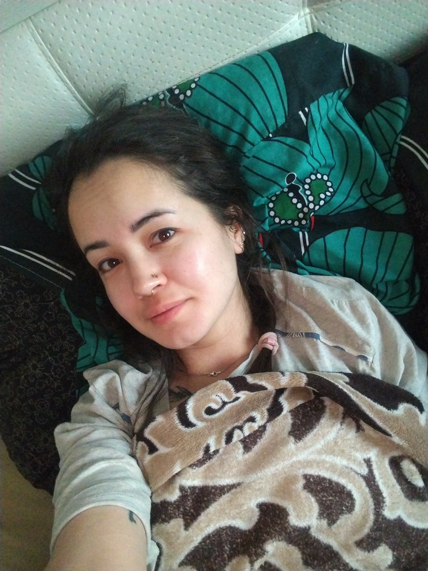 A person with dark hair and a nose piercing is lying on a bed. They are partially covered with a brown and white patterned blanket. The bed has a white headboard. Behind the person is a dark green blanket with a stylized design. The person is wearing a light gray long-sleeved shirt and a necklace. A small tattoo is visible on their left arm. The location is unspecified, and there is no evidence to suggest it is in the city of Noviy, Russia.