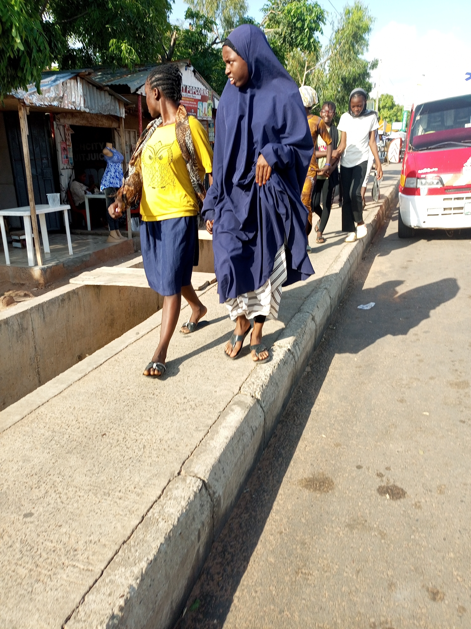 A medium shot captures a group of young women walking on a paved sidewalk next to a street in an urban area. The woman in the foreground, on the right, is wearing a navy blue hijab and a patterned skirt. She is walking away from the camera, her left side visible. To her left, another woman, partially obscured, wears a yellow t-shirt with an owl graphic and a blue skirt. She is also walking away from the camera. Further back, two more women are walking side-by-side. The woman on the right wears a white t-shirt and black pants, while the woman on the left has on a patterned brown and yellow outfit and glasses. Behind them, a red and white van is parked on the street. The environment appears to be a commercial area with small shops or stalls visible on the left, including one with "Y POPCORNS" signage. The ground is a mixture of concrete and asphalt, with a curb separating the sidewalk from the street. Trees and foliage are present in the background, suggesting an outdoor setting with daylight.