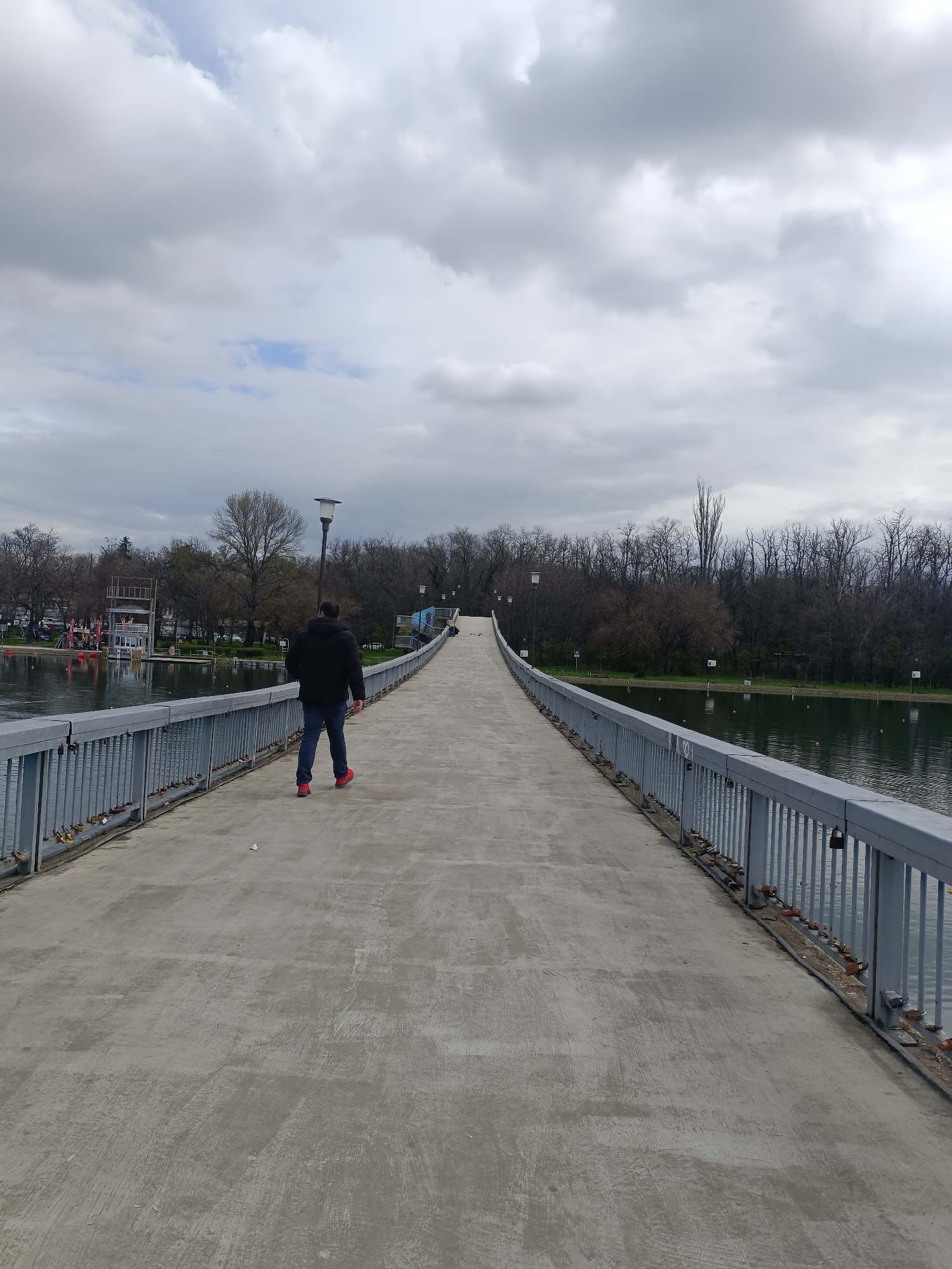 The image shows a man walking on a long, concrete pedestrian bridge. He is wearing a dark jacket, blue jeans, and red shoes, walking away from the camera. The bridge has metal railings on both sides and stretches over a body of water, likely a lake or river. On either side of the water are trees and greenery. In the background, a lifeguard tower can be seen. The sky is overcast with gray clouds. Love locks are fixed on the bridge. The location is the pedestrian bridge leading to the Rowing Channel in Plovdiv, Bulgaria.
