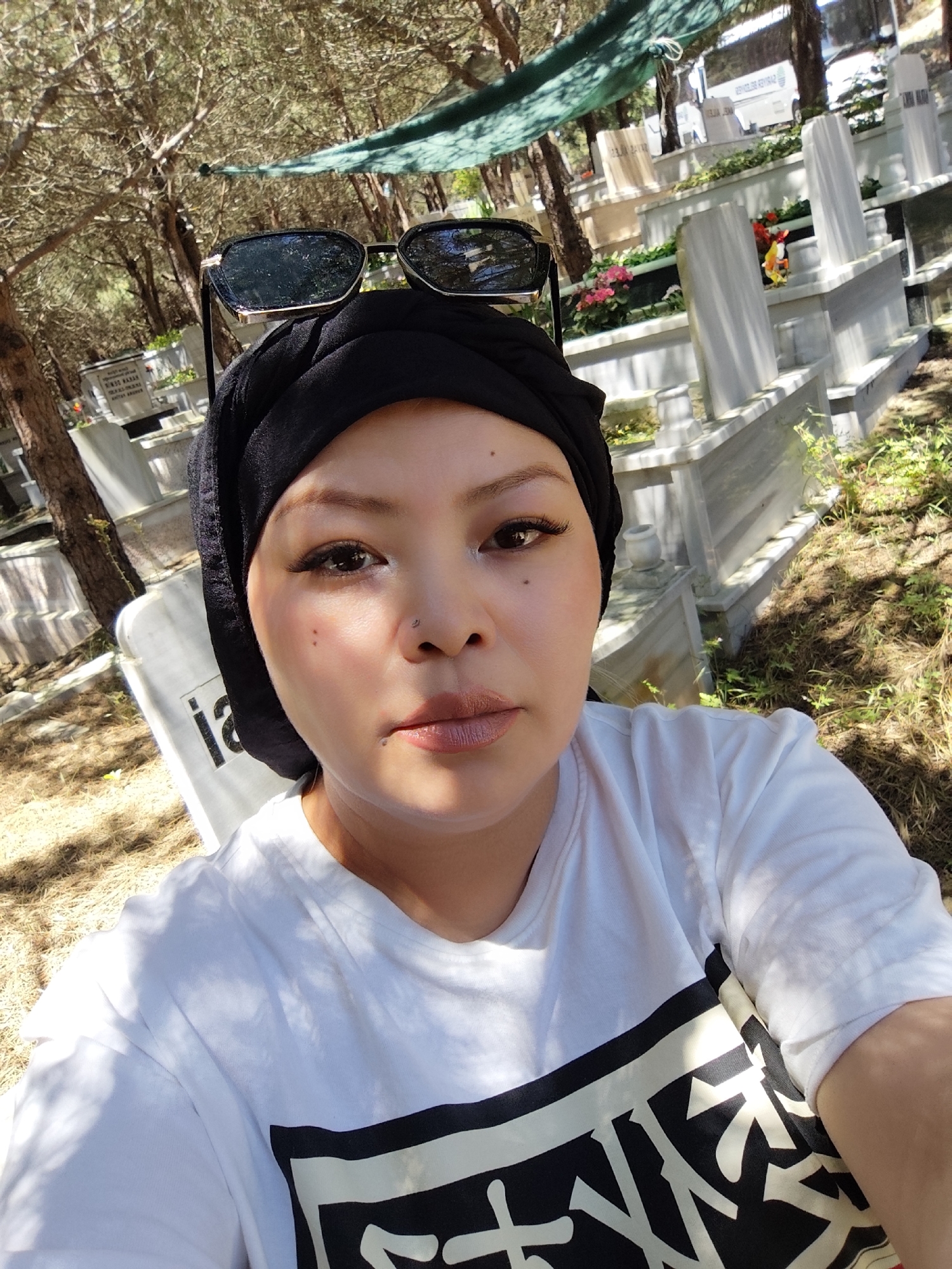 A selfie taken in a cemetery in Sarıyer, Turkey. The subject is wearing a black head covering and sunglasses on top of her head. She is wearing a white t-shirt with a black design on the front. In the background are rows of white gravestones. Trees and foliage are visible throughout the cemetery. The lighting suggests it is daytime.