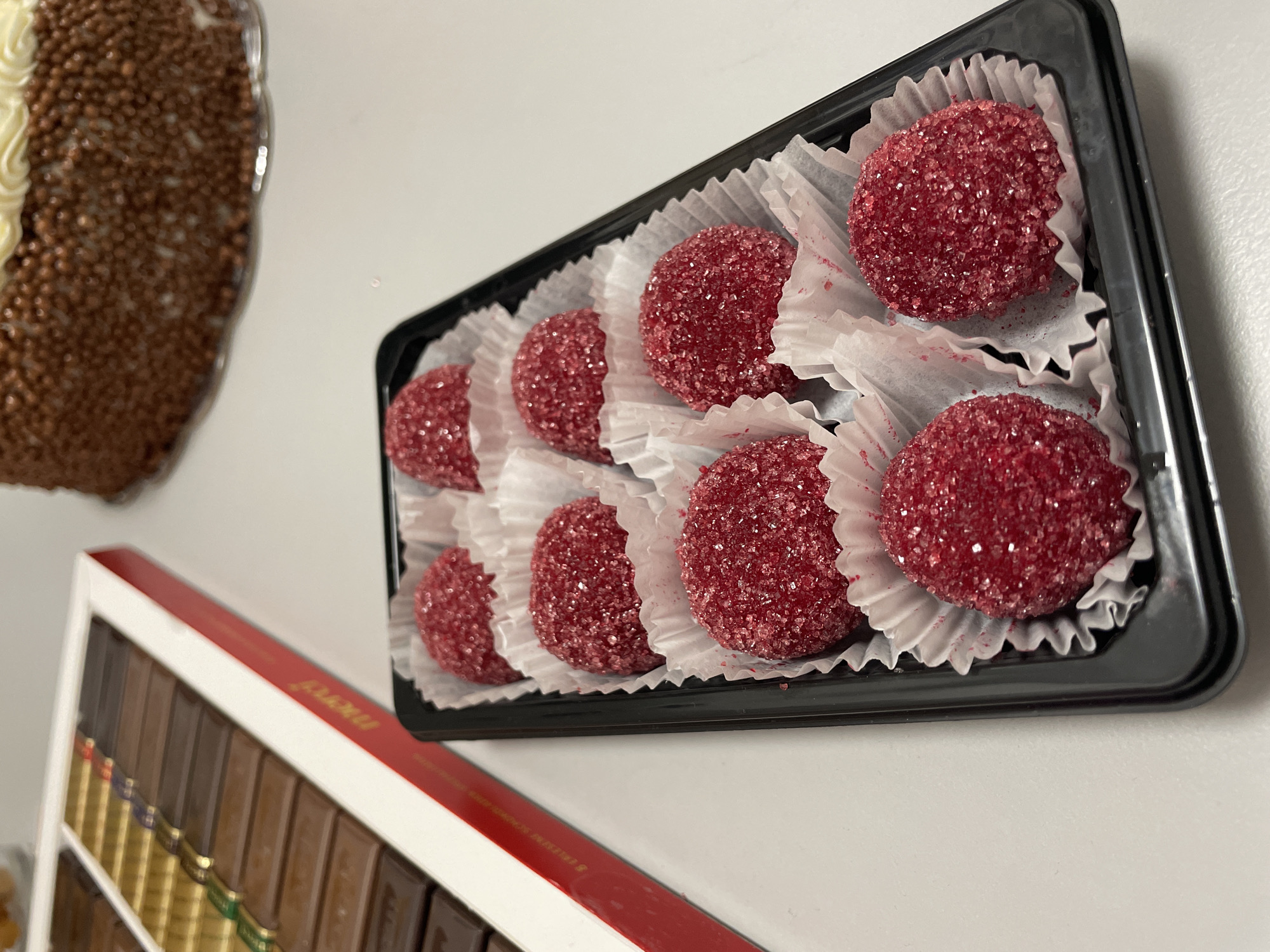 The scene contains a tray of eight red, sugar-coated candies in white paper cups, arranged in two rows within a black plastic tray. The tray is placed on a white surface. Adjacent to the tray is a partially visible box of assorted chocolates with a red and white border. In the background, slightly out of focus, is a cake decorated with chocolate sprinkles and white frosting. The location is likely Sofia, Bulgaria, indicated by the brand of chocolates, which is local to that area.