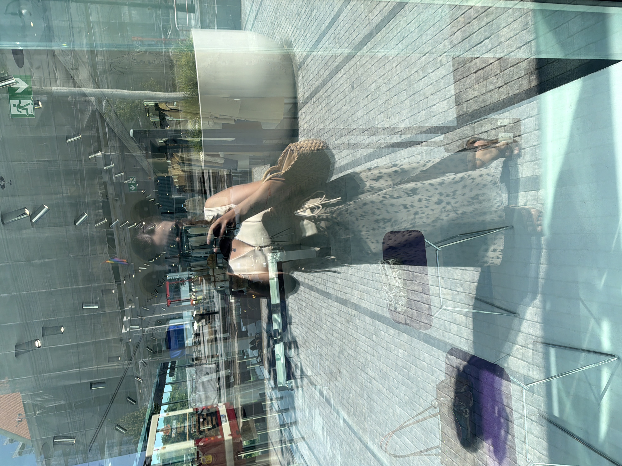 The image is a reflection on a glass storefront, likely in Prague, Czechia, showing a woman taking a selfie. She is wearing a light-colored, sleeveless top and a long, patterned skirt, along with sandals. A woven straw bag is slung over her shoulder. In the reflection, we see various elements of the storefront and its surroundings, including clothing racks, mannequins, and architectural details of the building, possibly in a shopping area. The ground outside appears to be paved with light-colored stones. The reflection creates a layered effect, with the interior of the store and the exterior environment merging together.