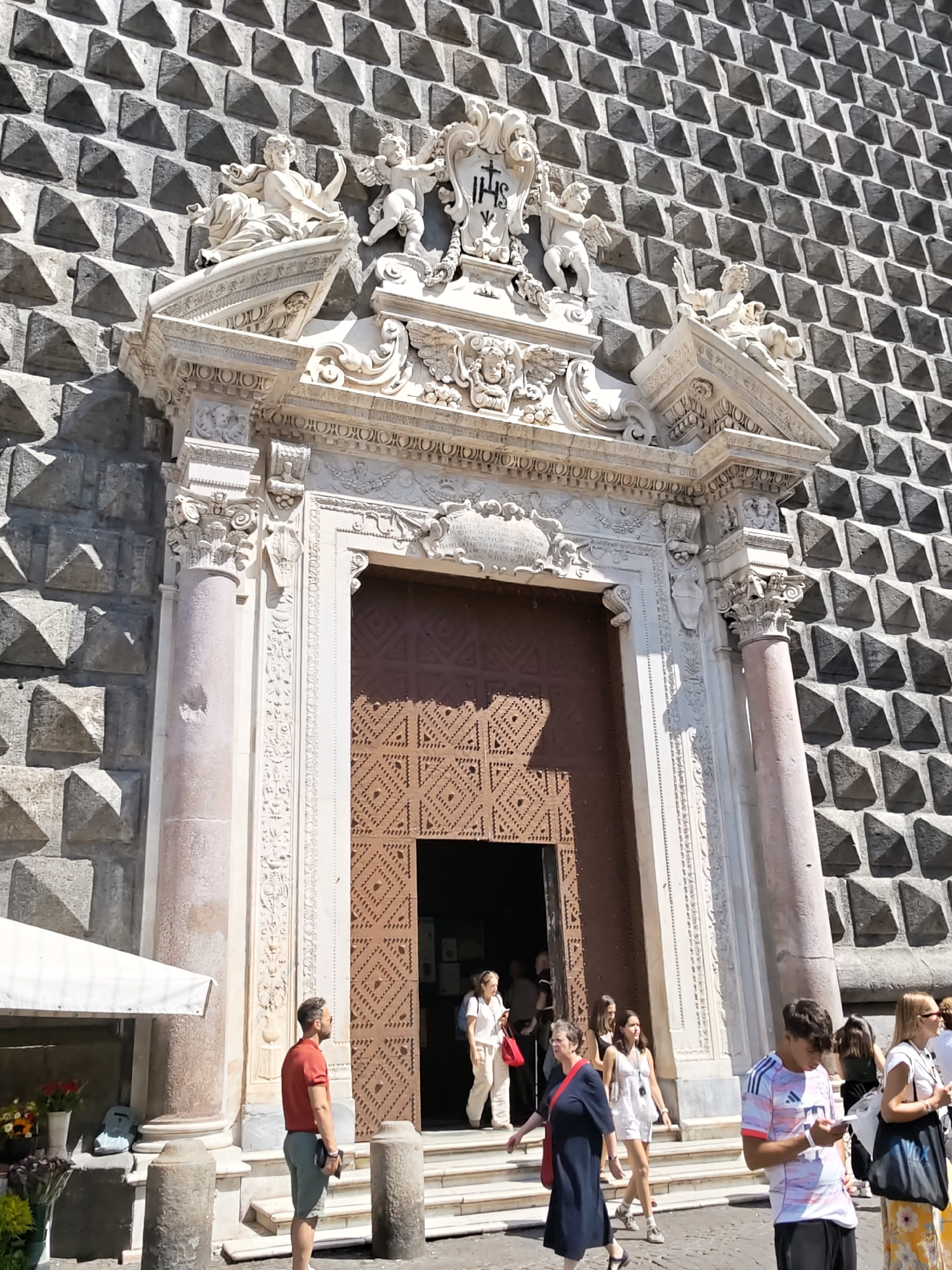 The image captures a sunlit exterior view of the Church of Gesú Nuovo in Naples, Italy, known for its distinctive diamond-faceted facade. The foreground features a few individuals; a man in a red polo shirt and grey shorts stands to the left, holding a phone. To the right, a young man in a sports-branded t-shirt is also looking at his phone. Several other people are seen on the steps leading up to the church's entrance, with a few entering and exiting the building. The ornate Baroque entrance is flanked by columns and adorned with sculptures. A white awning extends from a stall on the far left, with flowers displayed beneath it. The architecture of the church dominates the background, its geometric facade creating a textured visual pattern under the bright daylight.