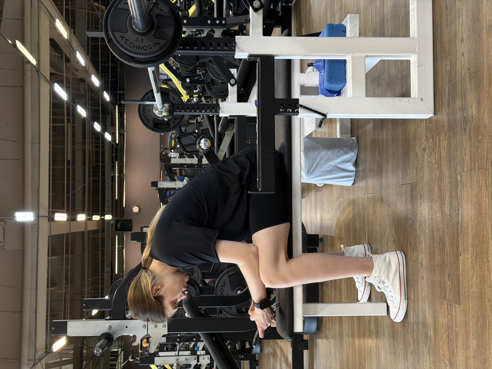 A young woman with blonde hair tied in a ponytail, wearing a black t-shirt and black shorts, is sitting on a weight bench in a gym. She is looking down at her phone, which she is holding in her left hand. Her right arm is resting on her bent leg. The bench is white and black, with black padding. There are weight plates and a barbell behind her, and other gym equipment is visible in the background. The floor is made of wood laminate. The lighting appears to be fluorescent. The image was taken in the city of Chaikovsky, Russia.