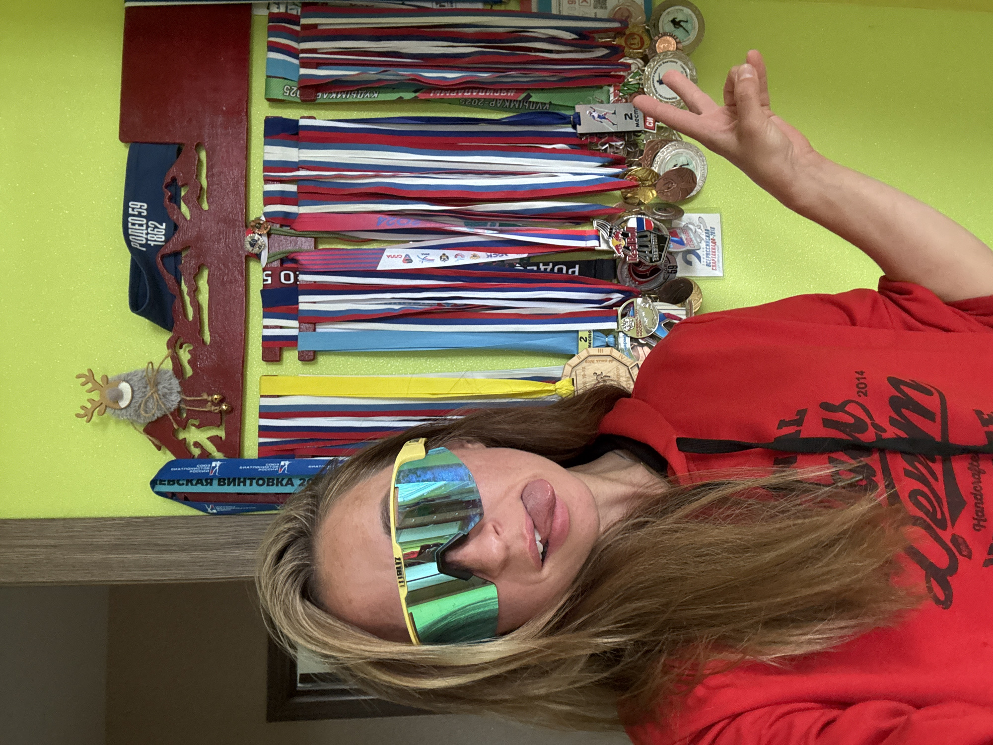 A woman, wearing a red hoodie and yellow sunglasses, sticks her tongue out and makes a peace sign with her fingers. She is in front of a wall displaying numerous medals, some of which are hanging from a wooden rack. The medals appear to be from various sporting events, with some ribbons featuring the Russian flag's colors. The room is brightly lit, possibly during the daytime. Visible text on the medals includes "РОДЕО 59 1862", "КУДЫМКАР-2025", and "ВСЕРОССИЙСКАЯ СПАРТАКИАДА-2018". The setting is indoors, with a wall painted a light green and a wooden shelf visible at the bottom. The woman's expression is playful and energetic.