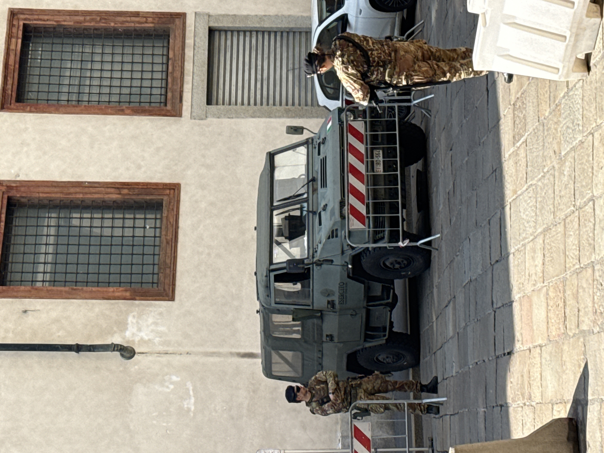 The image shows two Italian soldiers in camouflage fatigues standing guard next to a military vehicle in the city of Milan, Italy. The vehicle is a four-wheeled, light-green military truck with a ladder on the back. The soldiers are positioned at each end of the vehicle, near security barriers with red and white stripes. Behind the vehicle, there is a building with barred windows and a metal shutter. The location appears to be a public area with stone pavement. A white car is partially visible near the soldier at the top. The vehicle's license plate appears to read "E ESERCITO" followed by numbers.