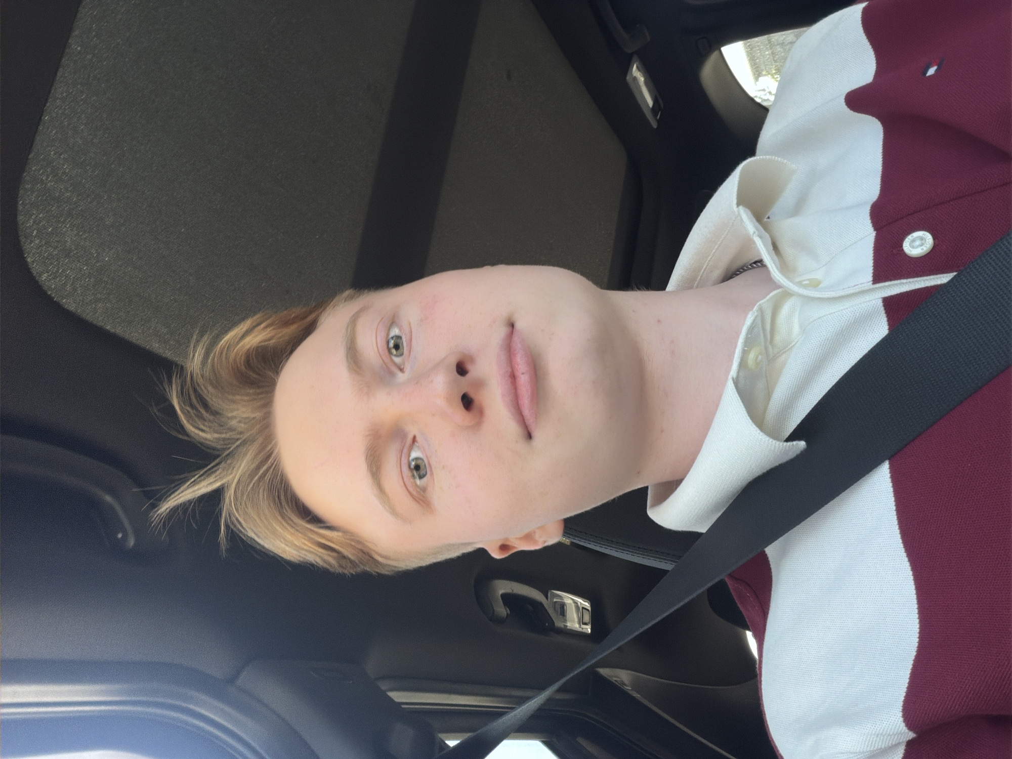 A young male with blond hair and fair skin is in the driver's seat of a car. He is wearing a collared shirt with horizontal bands of white and maroon. A black seatbelt is diagonally across his chest. The interior of the car, including the headliner and a portion of the dashboard, is visible. The background is out of focus and appears to be outdoors with daylight. The man is looking directly at the camera and has a neutral expression.