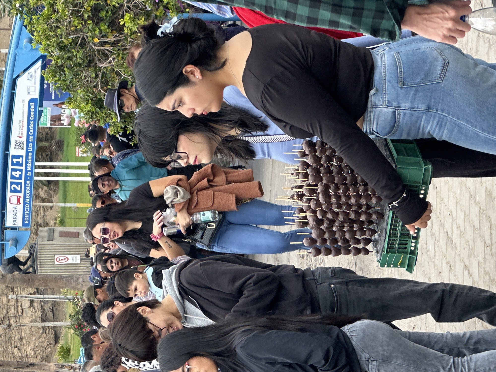 This outdoor image depicts a crowd of people in Arica, Chile, identified by a blue bus stop sign that reads "PARADA 2 4 1 EDIFICIO ALBORADA - Av Maximo Lira es Carlos Condell." The bus stop also displays text in Spanish about downloading a regional app and a message about city improvements. A vendor sells chocolate-covered strawberries on sticks from a green plastic crate. A woman in a black long-sleeved shirt and jeans stands holding the crate. Behind her, another woman in glasses and a brown jacket appears to be a customer. Various people are in the background, including a man in a hat and casual attire. The scene captures a public space, possibly along a street.