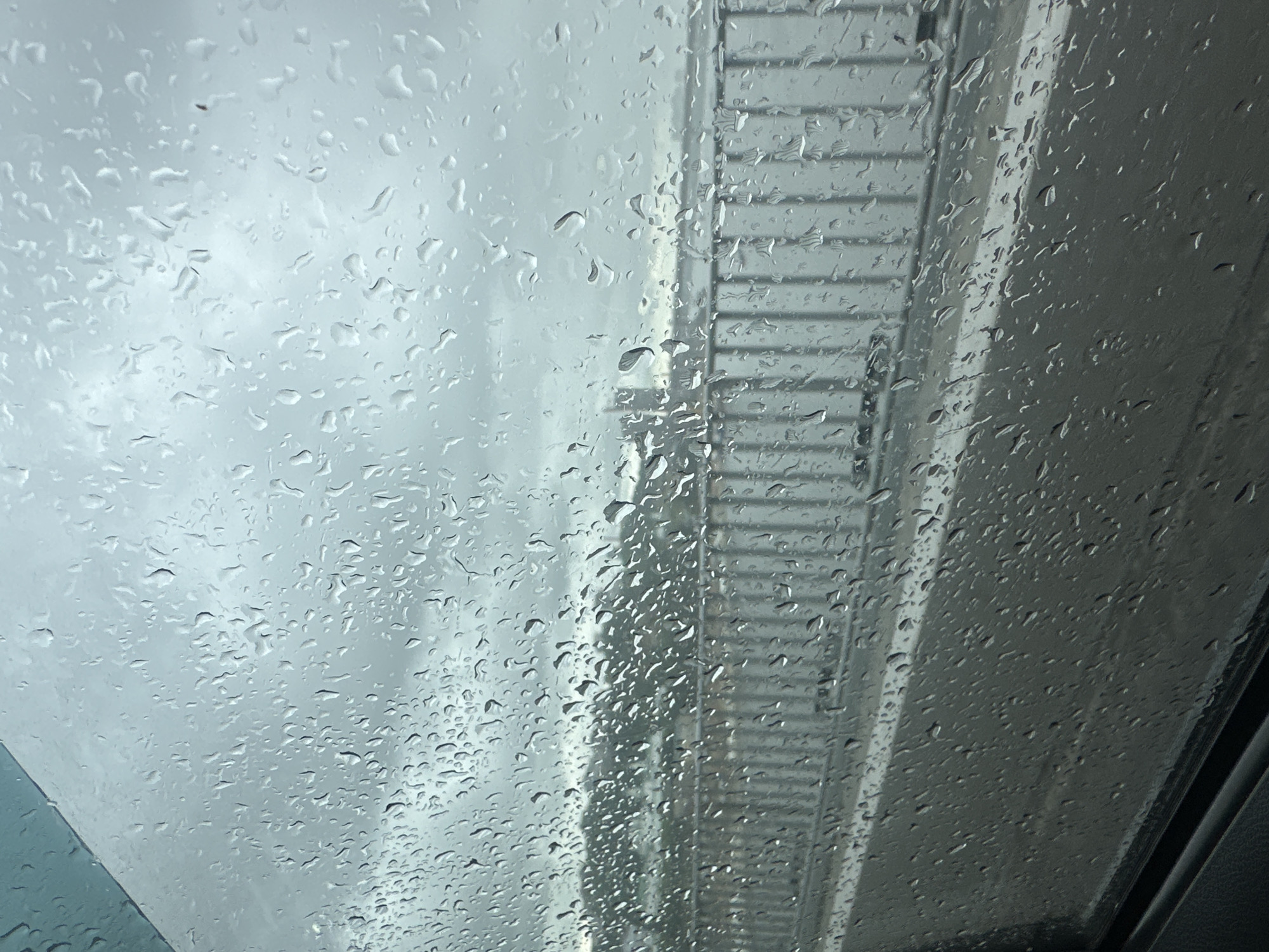 The image depicts a close-up view of raindrops on a window. The raindrops are varied in size and shape, distorting the view of the scene behind the glass. The background appears to be a cloudy sky, suggesting overcast weather or rain. To the right of the frame, there is a vertical structure with horizontal lines, possibly a building exterior or scaffolding, seen through the water-streaked glass. The colors are predominantly grayscale, with soft blues and grays. The overall mood is subdued, evoking a sense of being indoors during a rainy day. There is no visible text in the image, nor are there any people or discernible activities taking place. The time of day is suggested by the diffused light, which could be anytime during daylight hours when it's overcast. The location is not identifiable from the visual cues.