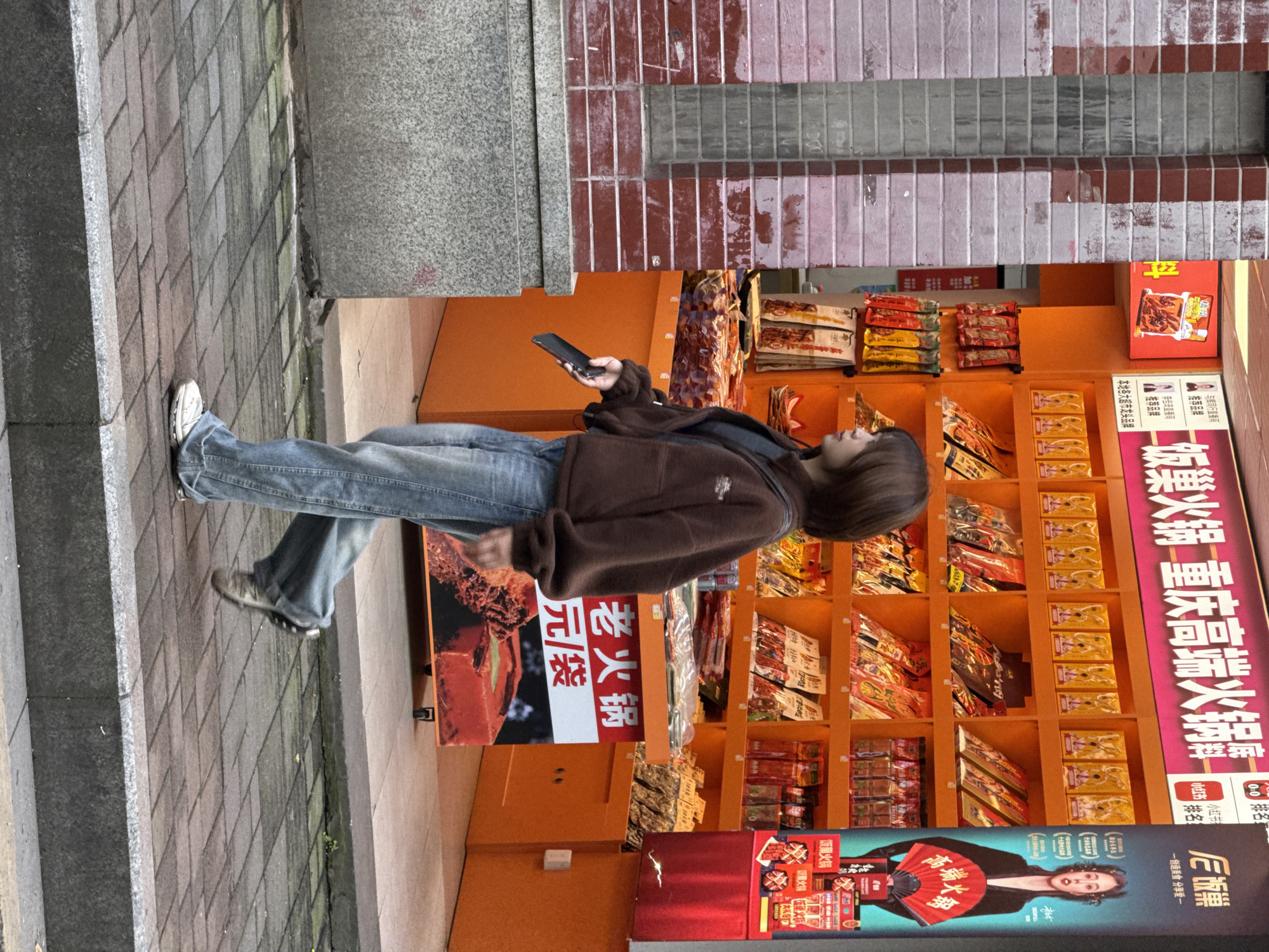 Here is a forensic description of the image.
The photograph appears to have been taken in the Yuzhong District of China, as evidenced by the Chinese characters on the signage. A person is standing to the side of an orange shelving unit, possibly located outside a shop or restaurant. The shelving is filled with a variety of food products.
The individual, possibly female, is wearing blue jeans and a brown jacket. She is holding a smartphone in her right hand, suggesting she may be checking her phone or taking a photograph.
The signage on the shelves features Chinese characters and promotional materials for a product called "饭巢火锅 重庆高端火锅", which translates to "Rice Nest Hot Pot Chongqing High-End Hot Pot." Other signs mention "老火锅" (Old Hot Pot) and "元/袋" (Yuan/Bag), indicating the price or serving size.
The surrounding environment includes a paved walkway and a brick wall, suggesting an urban setting. Overall, the scene is suggestive of a busy commercial area where people might stop to browse or purchase food items.
