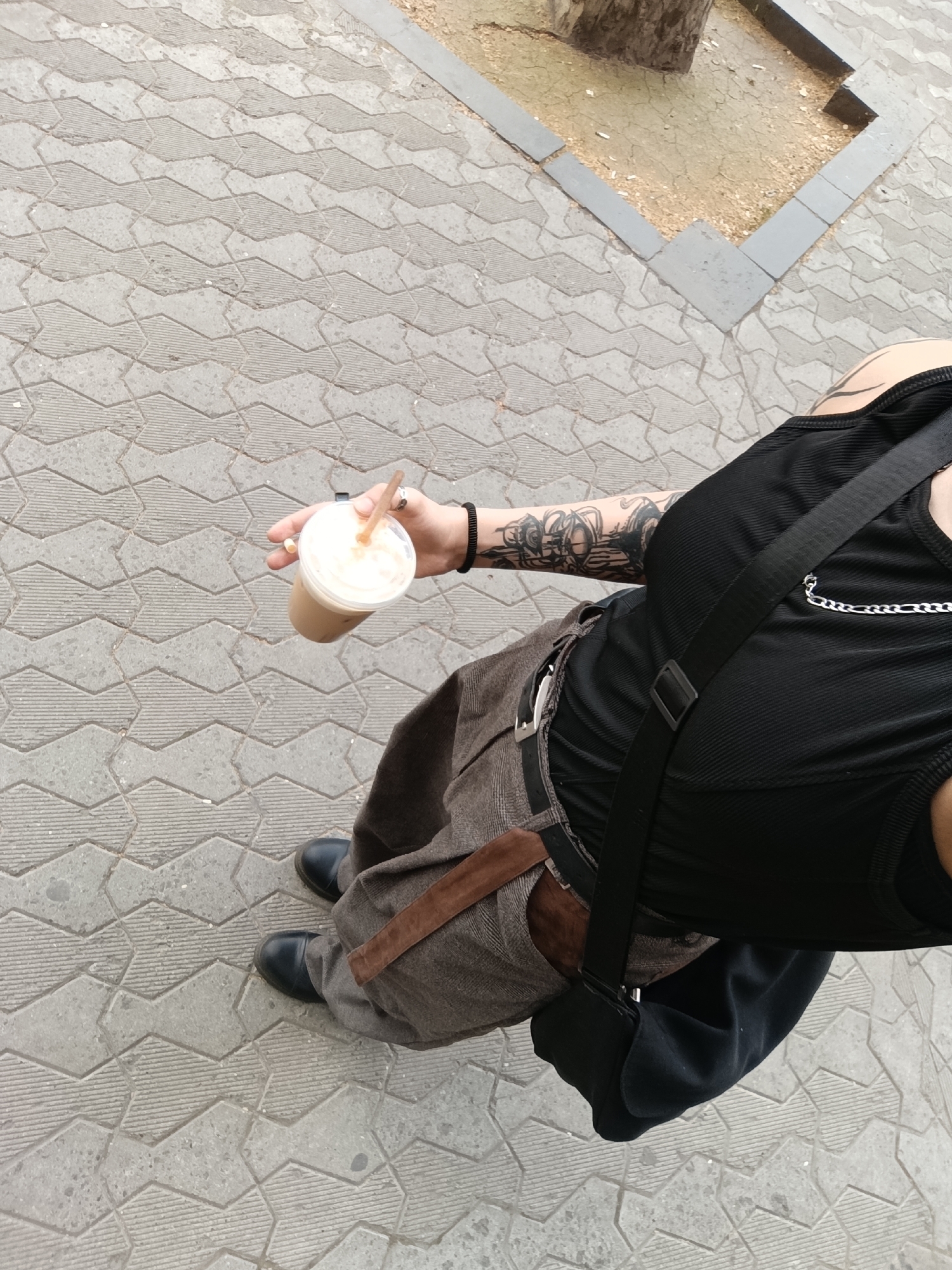 A downward-angled eye-level shot captures a person in Yerevan, Armenia. They are holding an iced coffee. The person is wearing a black tank top with a silver chain accent, brown trousers with a brown belt, and black shoes. A black bag strap crosses their chest. The person has tattoos on their arm. The ground is paved with gray interlocking stones. In the background, there's a tree planted in a bordered section filled with dirt.