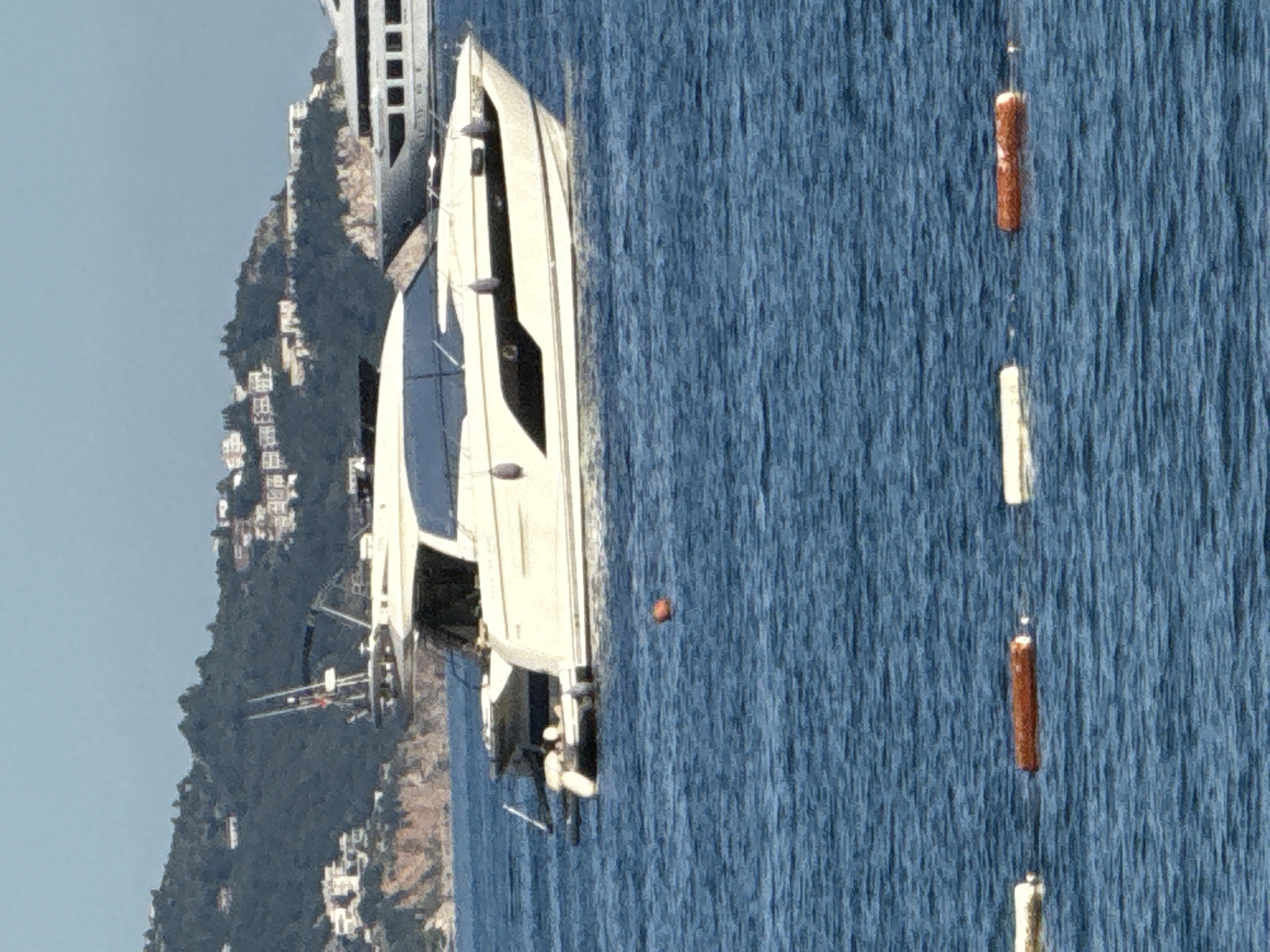 This image depicts a marine scene in Budva, Montenegro, under clear daylight conditions. The foreground features choppy blue water with an orange spherical buoy to the left of the image center and a horizontal line of three rectangular buoys (two white, one reddish-brown) closer to the bottom edge. The primary subject is a white motor yacht, positioned slightly right of center, with a dark-tinted cabin structure, numerous black fenders hanging from its starboard side, and a tender partially visible at its stern. A mast with antennas and equipment is visible on the yacht's upper deck. In the mid-ground, a larger, dark-hulled superyacht is partially visible to the right, showing multiple decks and windows. The background is dominated by a verdant, tree-covered hillside, populated with numerous white residential buildings clustered across its slopes. A rugged, light-colored cliff face is visible at the base of the hill where it meets the sea. No individuals are discernibly present.
