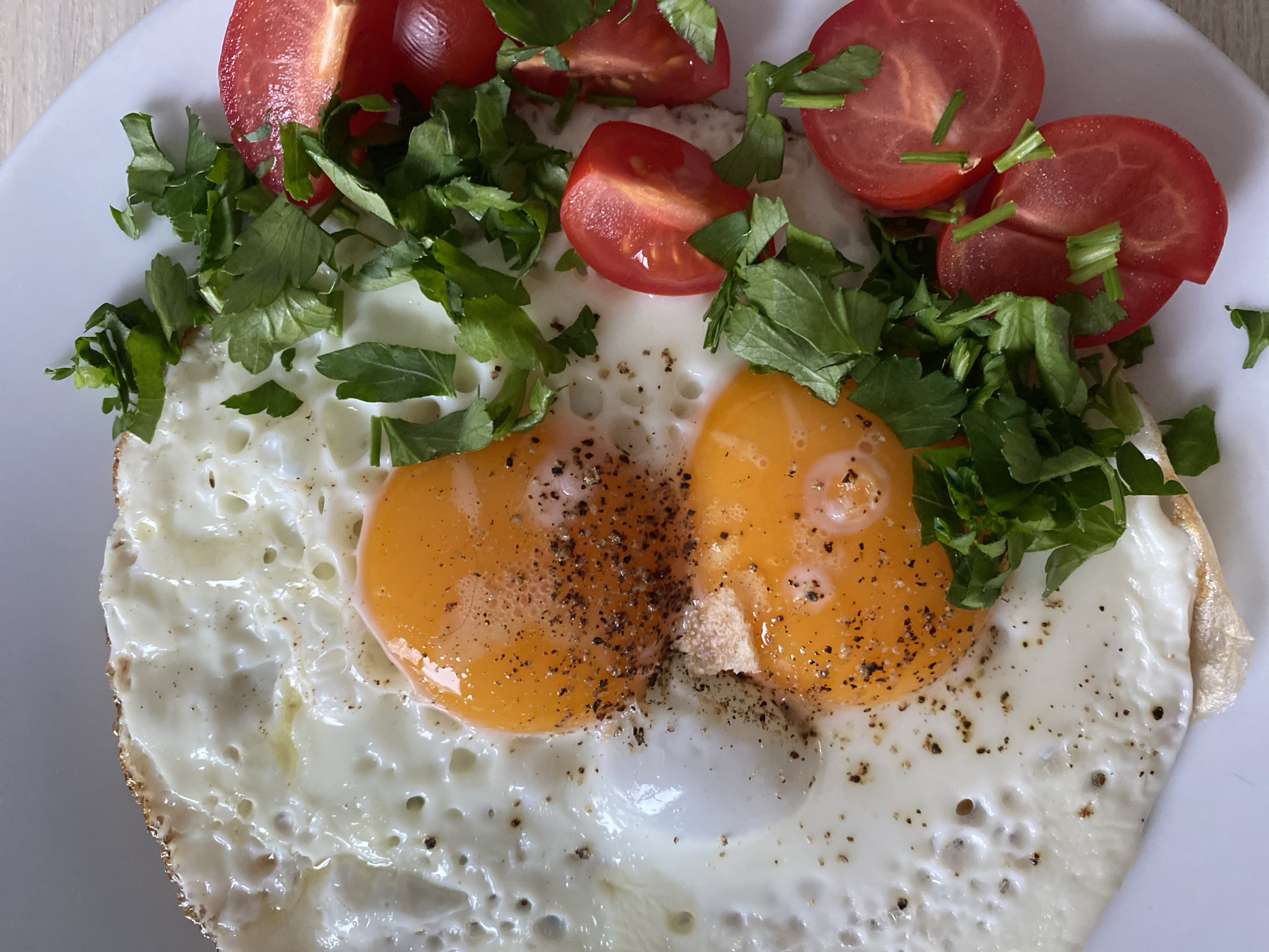 A close-up photograph shows a plate of food. Two fried eggs with yolks intact are centrally located, seasoned with black pepper. Chopped parsley and quartered cherry tomatoes are arranged on top and around the eggs. The plate is white. The image does not provide enough information to establish the location as Chaikovsky, Russia.