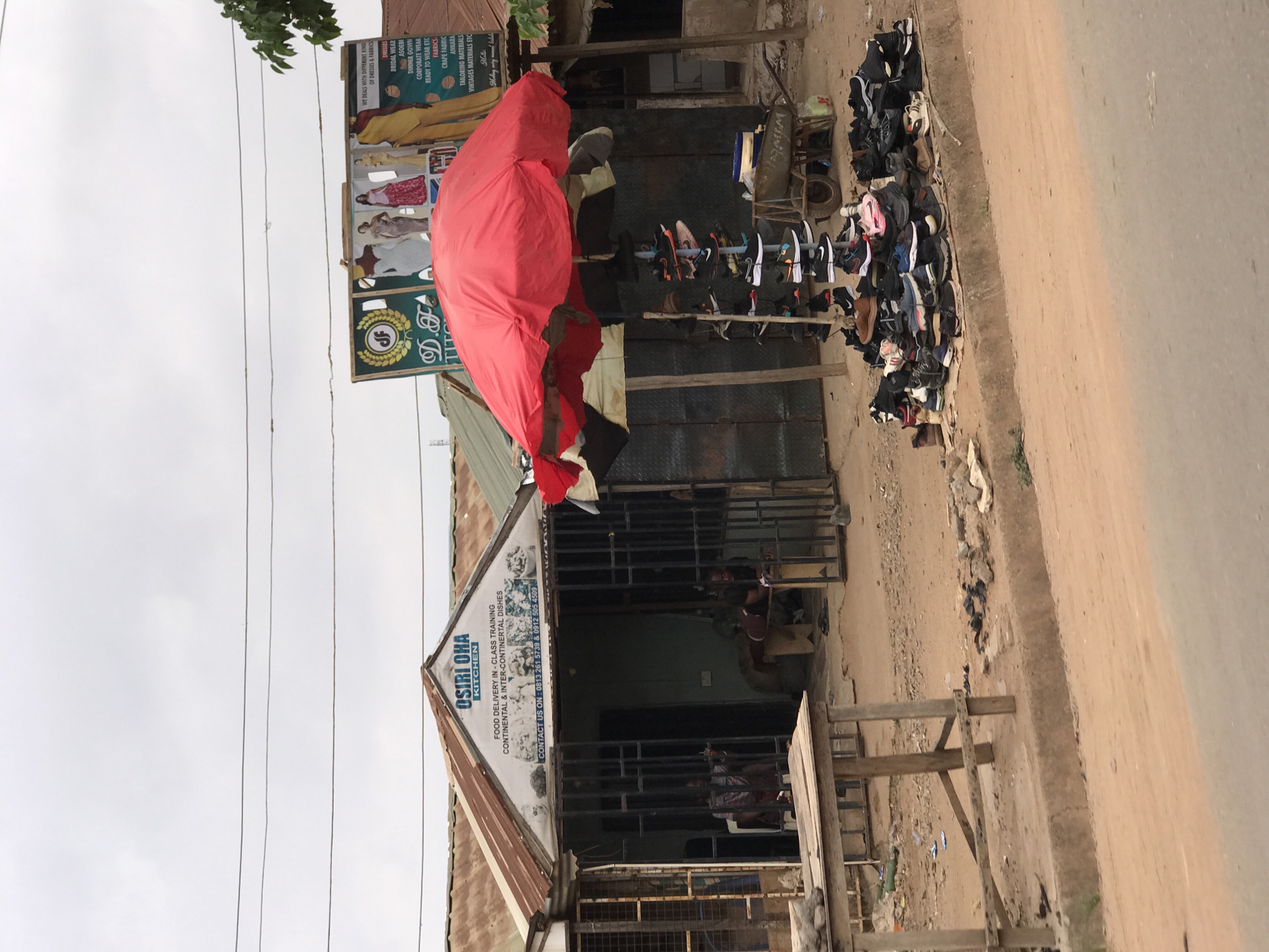 The image depicts a street scene in what appears to be a commercial area of Abuja, Nigeria. A storefront identified as "OSIRI OHA KITCHEN" is visible, suggesting a food delivery or training business. Next to it is another establishment "D.T. D" selling dresses and fabrics, indicated by signage. A red cloth or awning is draped over a portion of the front area. Shoes are displayed for sale on racks along the side of the road. People are seated or standing inside the storefronts. The surrounding environment shows a partially paved road with dirt or gravel edges. The sky is overcast.