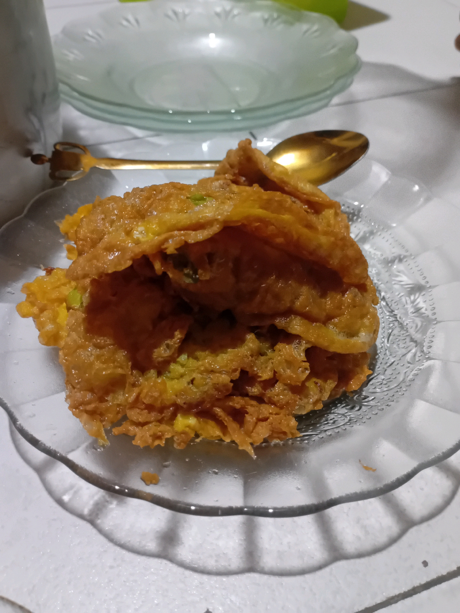 A close-up shot features a golden-brown omelet, appearing crispy and folded, served on a clear glass plate with a decorative rim. The plate rests on a white, textured surface, possibly a tablecloth. To the left, a portion of a white, marble-patterned object, possibly a container, is visible, and a brass-colored utensil with a distinctive handle is placed horizontally behind the omelet. In the background, several stacked clear glass plates are visible, one with a subtle green tint. The lighting creates shadows on the white surface, indicating an indoor setting. This scene likely depicts a meal in Pekanbaru, Indonesia, given the cuisine style.