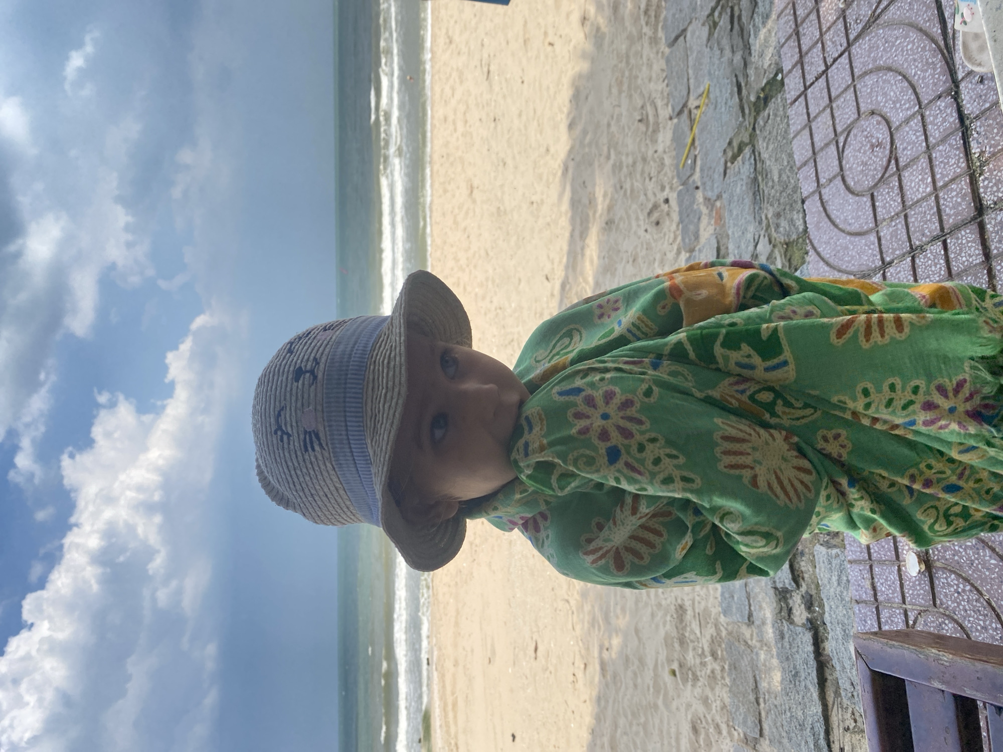 A young child wearing a straw hat and a green patterned sarong stands on a paved area near a sandy beach in Mũi Né, Vietnam. The child is looking down and to the side, with their hands near their face. The beach is visible in the background, with gentle waves rolling in from the sea under a cloudy sky. The paved area has a patterned design and is adjacent to a stone retaining wall and a wooden bench.