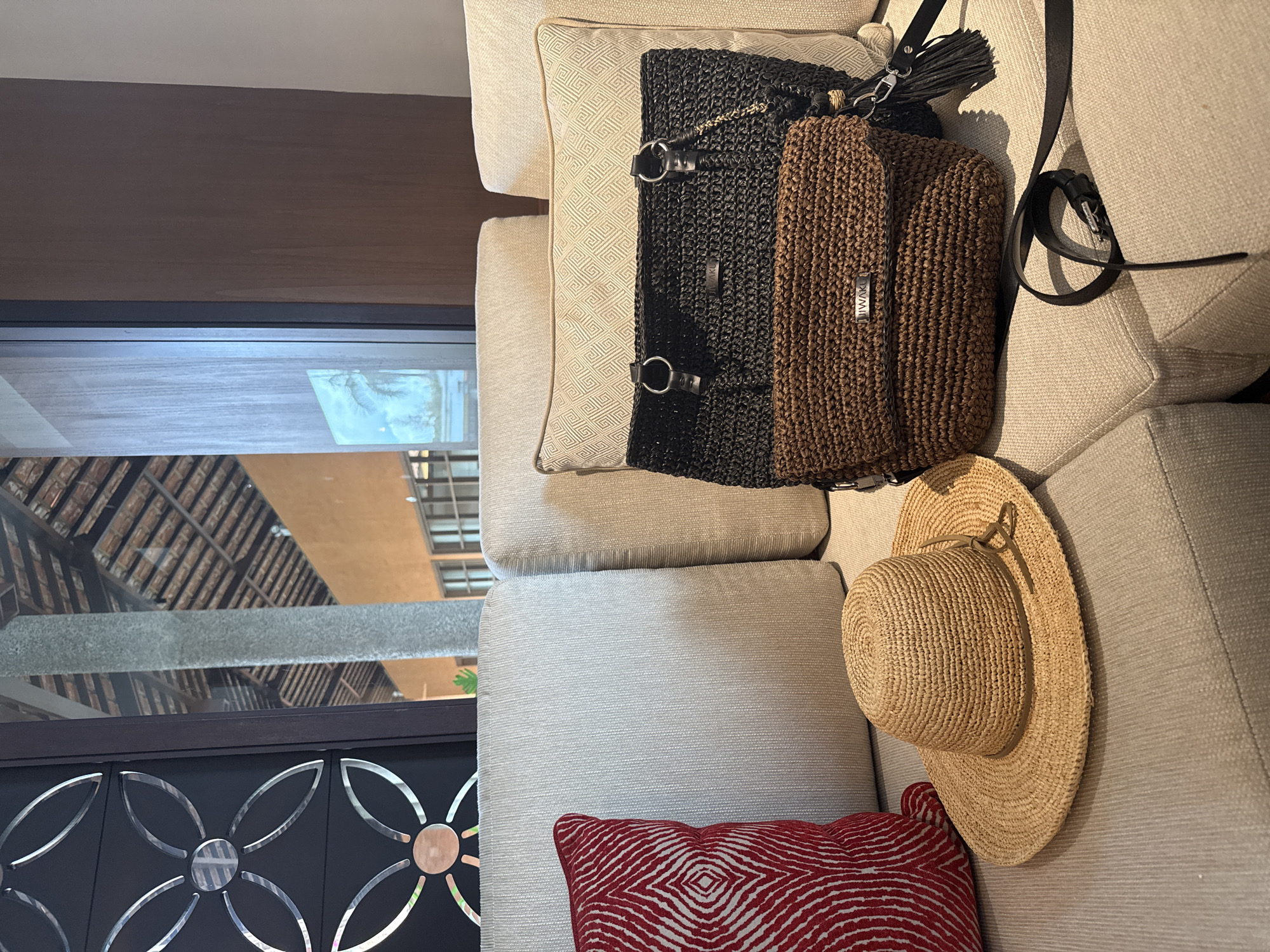 A straw fedora hat is positioned on a tan sofa next to a red and white patterned pillow. To the right of the hat, two woven handbags, one black and one brown, are placed on the sofa, with a black leather strap extending across the sofa in the foreground. The scene is set indoors, likely in a lounge area, with a large window in the background revealing a view of an outdoor space with a tiled roof structure and a stone pillar, possibly indicating a resort or hotel in a tropical location such as Kuta Selatan, Indonesia.