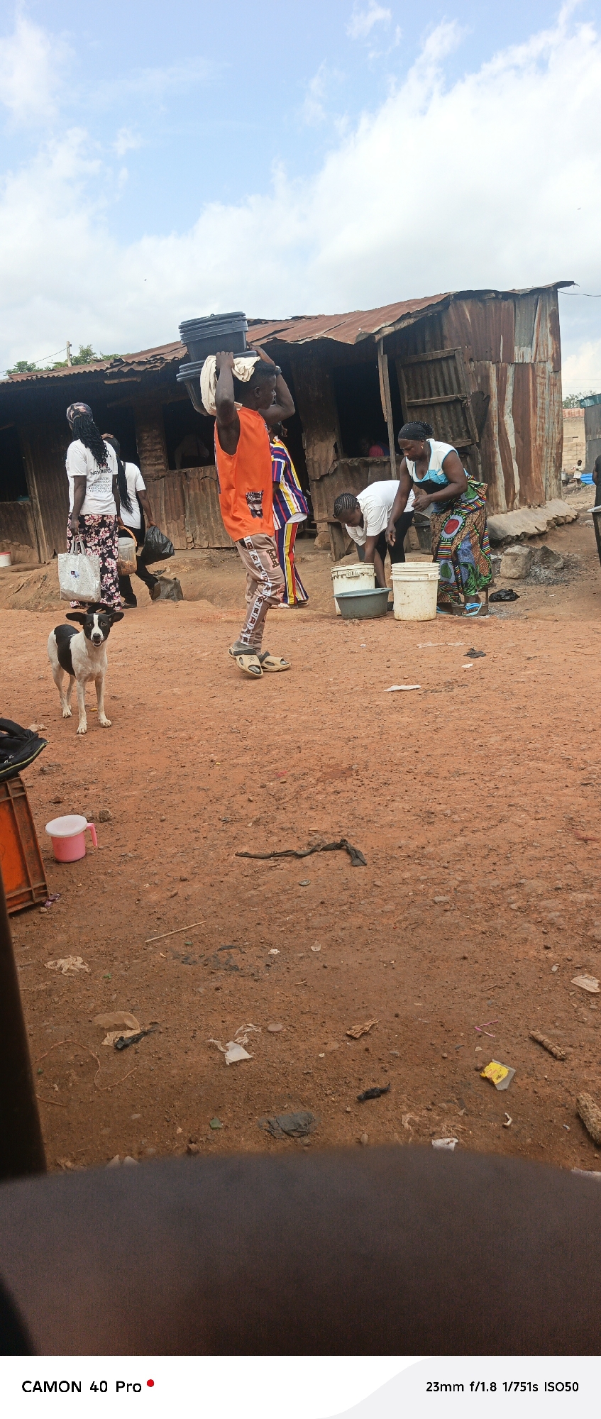 In Barikin Gidar Kanat, Nigeria, a man carries several black plastic containers on his head, balanced with the help of a white cloth. He is dressed in an orange sleeveless shirt and patterned pants, with light-colored sandals on his feet. Beside him, a woman in a white and blue top and a colorful patterned skirt is attending to buckets on the ground. Another woman, wearing a white top and a patterned skirt, is visible in the background, holding a shopping bag. A black and white dog stands alert in the foreground, looking towards the camera.
The scene appears to be an outdoor setting with a rustic, possibly impoverished, dwelling in the background, constructed from corrugated metal and wood. The ground is covered in reddish-brown dirt, with scattered debris. To the left of the frame, an orange plastic crate and a pink cup are visible.
The sky above is partly cloudy, suggesting daytime. The overall impression is one of daily life and resourcefulness in a rural Nigerian community. The visible text on the image reads "CAMON 40 Pro" and "23mm f/1.8 1/751s ISO50", indicating the camera model and settings used to capture the photograph.
