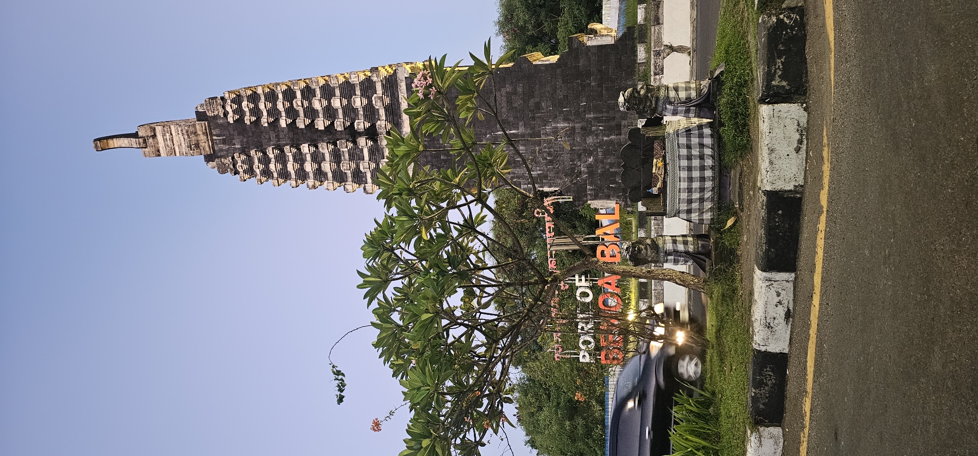 The image shows a view in Kuta, Indonesia. On the left side of the image is a towering, stepped temple structure made of dark stone. In front of the temple, a tree with green leaves partially obscures an orange sign that reads "PORT OF BENOA BALI." Two statues draped with checkered cloth are placed in front of the sign. A car with its headlights on is visible behind the sign. The scene is situated along a road with a black and white curb and a yellow line separating the lanes. Grass grows between the road and the monument area. The sky above is clear and light blue.