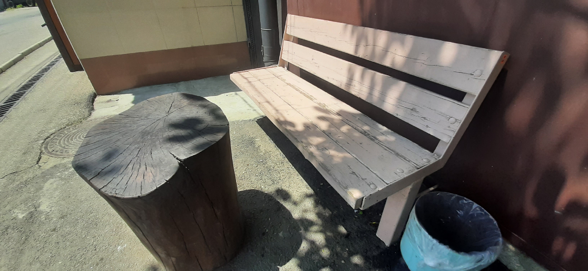 A light-colored, wooden slatted bench with peeling paint is positioned against a dark brown wall. To the left of the bench is a large, dark-colored wooden stump serving as a seat. In front of the bench, to the right, is a light blue trash can lined with a white plastic bag. The scene appears to be outdoors, with a concrete or asphalt ground surface, a metal grate drain, and a manhole cover visible to the left. Sunlight casts shadows of tree leaves onto the bench and ground. The image is taken on a sunny day.