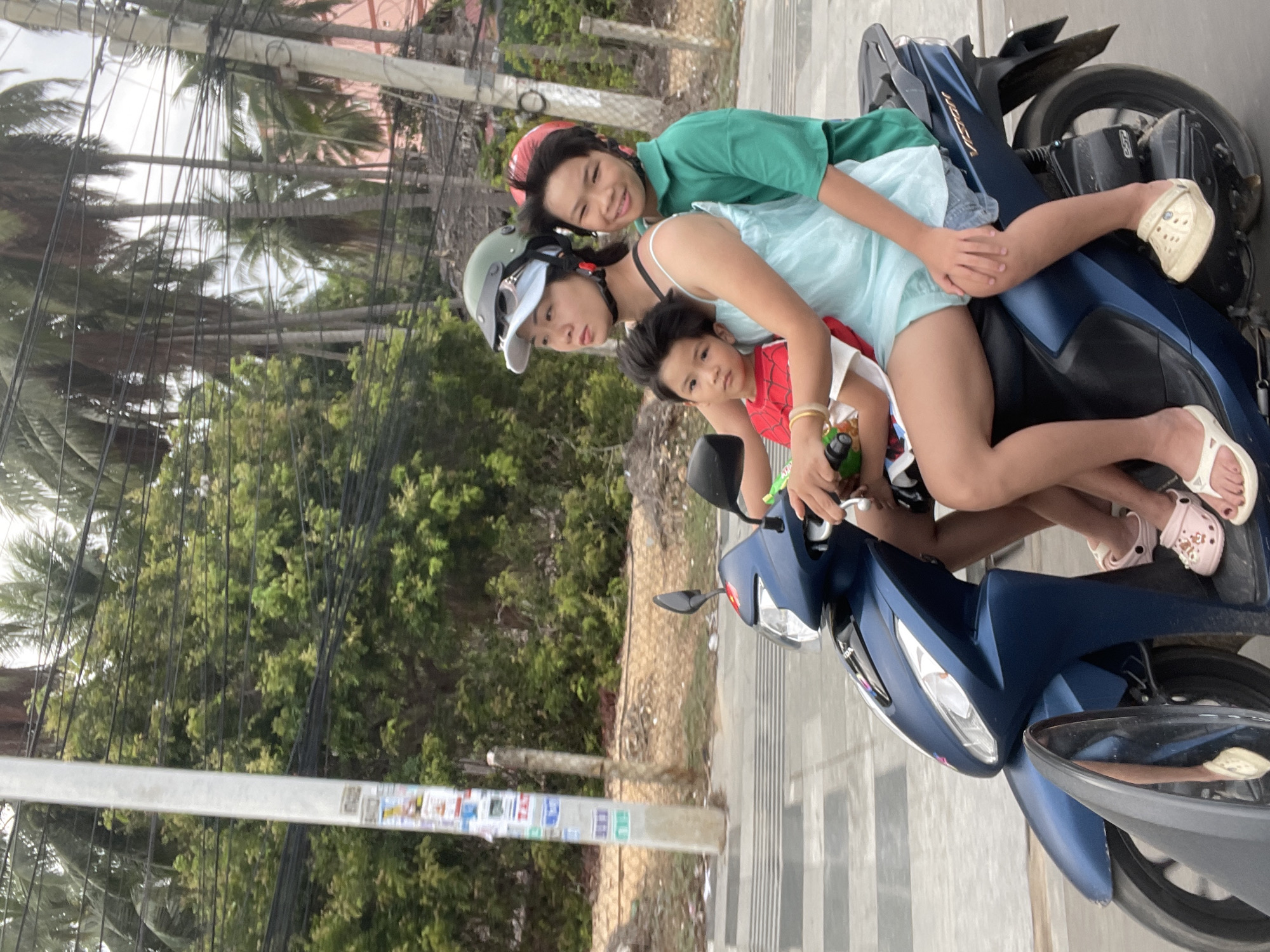 Three people are on a blue scooter. A woman in a light blue dress and a gray helmet is in the middle. A child wearing a Spider-Man tank top is in front of her, seemingly at the helm of the scooter. A person in a green shirt and a red helmet sits on the back of the scooter. They are on a paved road or sidewalk. The background includes trees, utility poles, and buildings. The location is Phu Thuy Ward, Vietnam.