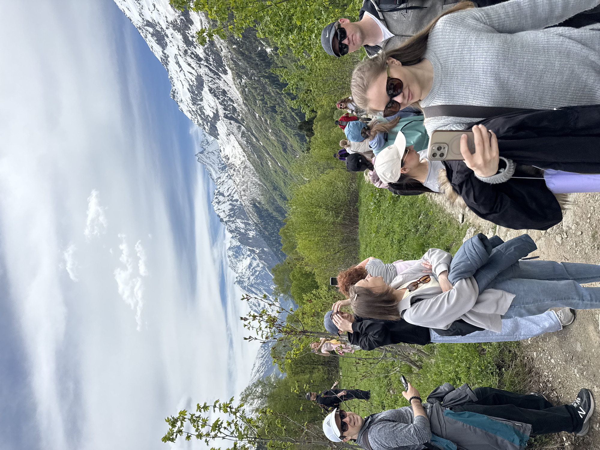 The image depicts a group of approximately 15-20 individuals on a hiking trail in a mountainous region of Верхне-Тебердинское сельское поселение, Russia. The scene is set under a partly cloudy sky with prominent snow-capped mountains dominating the background. Lush green trees and bushes, many with vibrant new leaves, line the dirt path that recedes into the distance.
In the immediate foreground, a young woman with long brown hair and sunglasses, wearing a light grey knitted sweater, is visible on the right. She holds a gold-colored smartphone horizontally in her right hand, aiming it towards the left, possibly taking a photo or video. A black shoulder strap crosses her chest, and she carries a black jacket over her left arm. To her right, a man in a dark grey hooded top, sunglasses, and a black baseball cap is partially visible, looking towards the right. On the left foreground, a man wearing a white baseball cap, sunglasses, and a grey long-sleeved shirt with a backpack is looking directly at the camera, holding a dark smartphone in his left hand. A grey jacket is tied around his waist, and he wears black New Balance sneakers.
Further back on the left, a woman in a light grey jacket and blue jeans stands with her arms crossed, holding a blue jacket. Adjacent to her, a young man in a dark hoodie and a blue baseball cap is seen adjusting his hair. Behind them, a woman with reddish-brown curly hair in a striped top looks towards the mountains. Another woman in a pink top is visible in the midground, holding a phone to capture the scenery. A man in black attire and a black baseball cap stands further back on the left. Several other individuals are dispersed along the winding dirt path, walking away or paused, dressed in various casual outdoor clothing, including jackets, caps, and sunglasses. The overall impression is of a group engaging in outdoor recreation amidst a striking natural landscape.