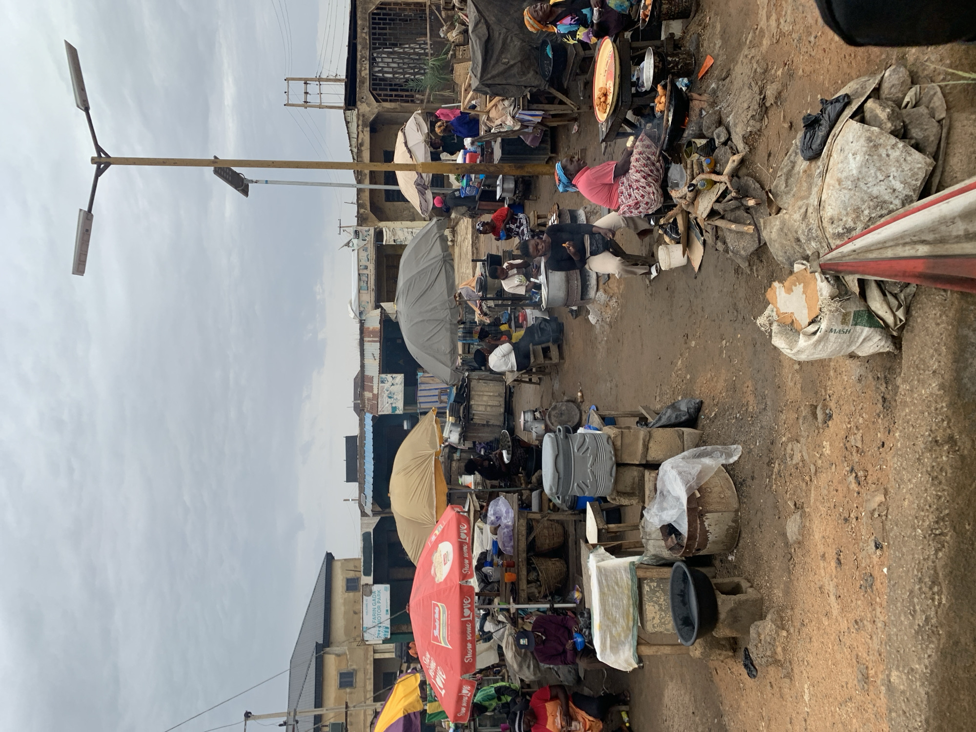 The image shows a bustling marketplace in Rafin Par, Nigeria. The market is located on a dirt street under a cloudy sky. People are buying and selling goods from makeshift stalls and tables, many of which are shaded by umbrellas. Stalls are cluttered with a variety of objects including cooking pots, baskets, and packaged goods. Some people are seated, possibly cooking or selling food, while others are moving through the crowded space. A lamppost is visible in the foreground. The environment is dusty and somewhat chaotic. Buildings line the street in the background.