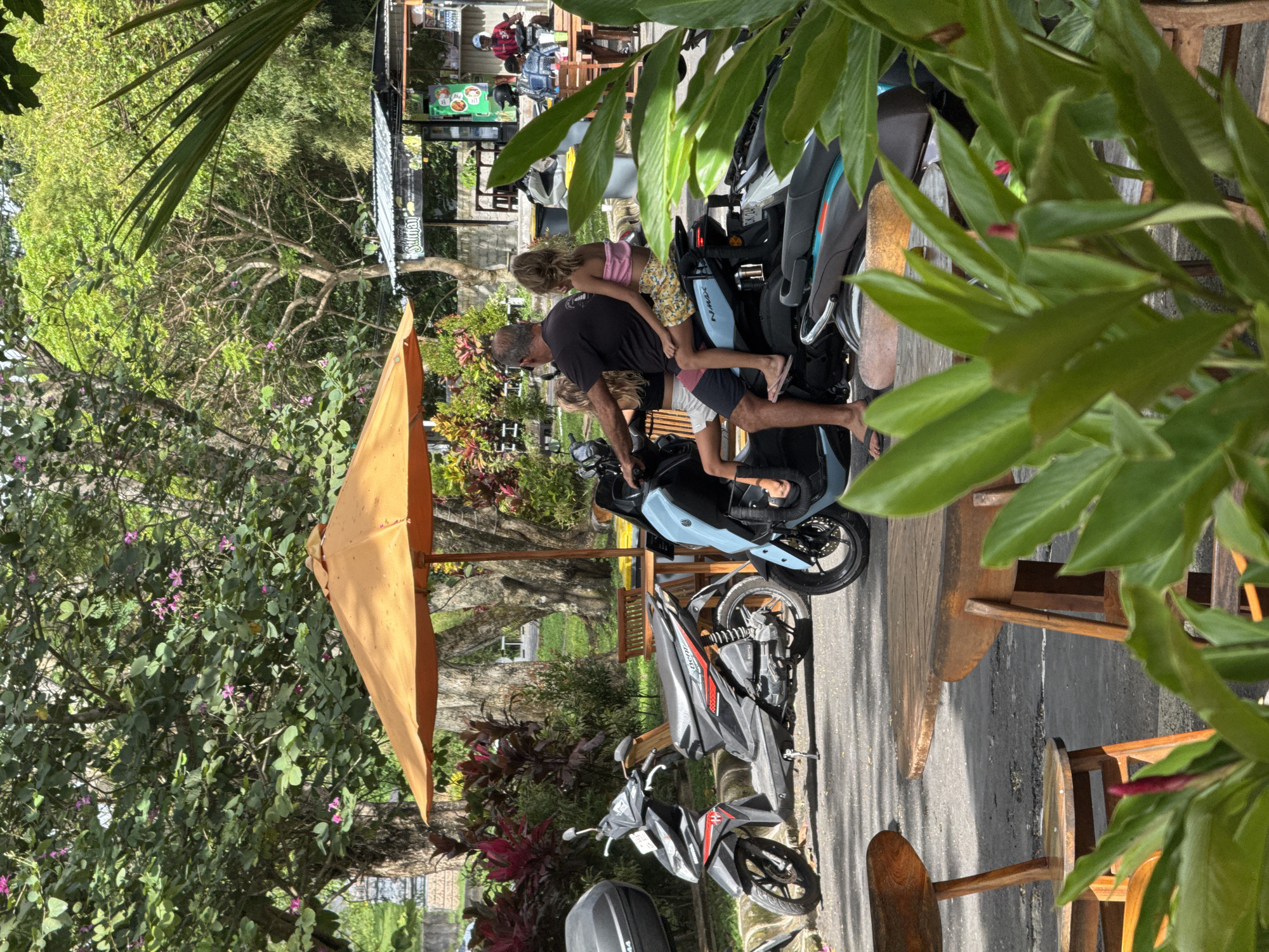 Captured at an outdoor setting in Kuta Selatan, Indonesia, the image shows a man with two children near parked scooters. The man, wearing a dark shirt and light shorts, is standing beside a light blue scooter. Two children, one wearing a pink top and floral shorts, the other wearing a white top, are beside him on the scooter. Adjacent to the blue scooter is another parked motorcycle and a partially visible gray scooter. Further in the background is a seated group of people under a structure, seemingly an open-air cafe or vendor. The scene is framed by foliage, including an orange umbrella and tropical plants. Wooden tables and chairs are also visible, reinforcing the outdoor cafe setting.
