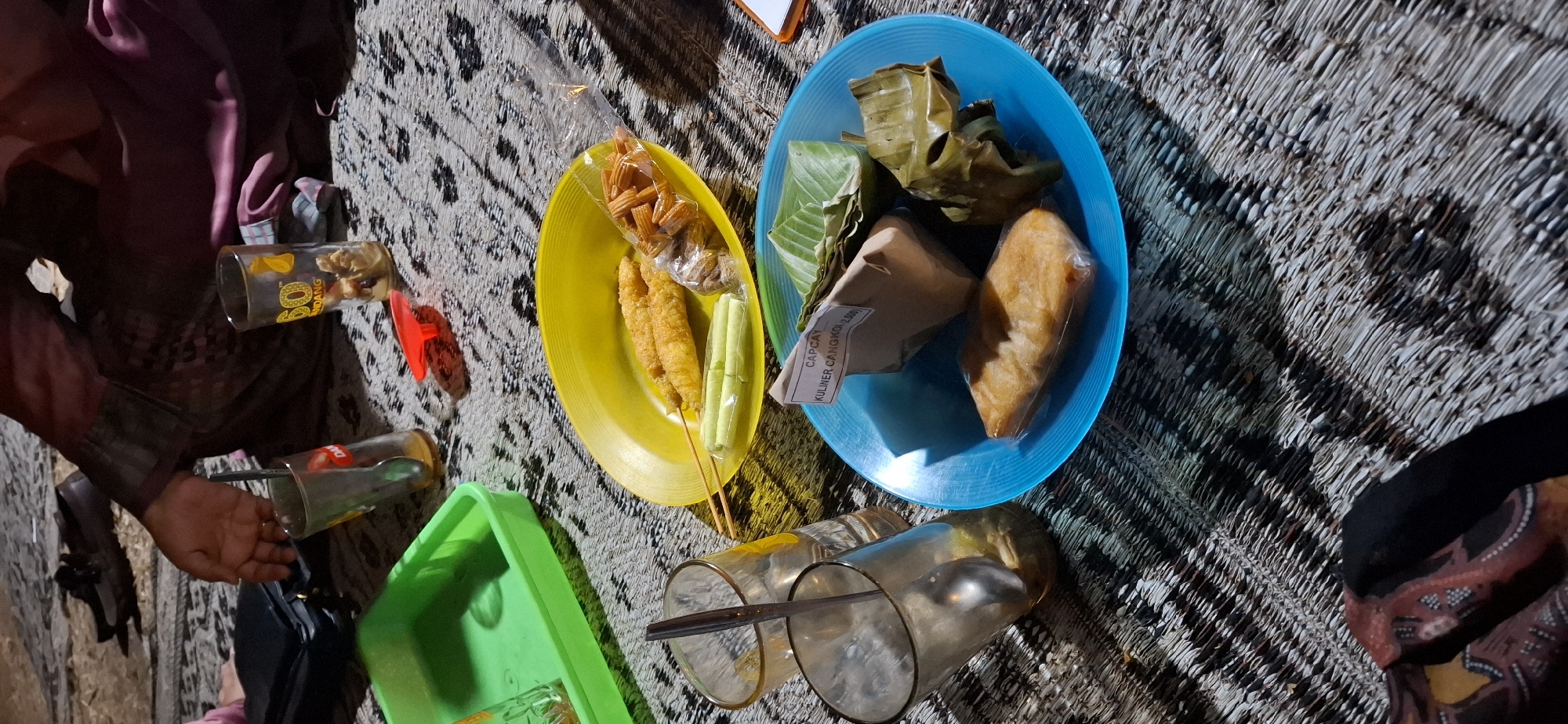 The image depicts a low-angle, close-up view of food and drinks arranged on a woven mat, likely on the ground, in Cangkol, Indonesia. The setting appears to be casual, possibly an outdoor gathering or a picnic.
In the foreground, a blue plastic plate holds several food items. There is a piece of fried food, possibly fish or a fritter, in plastic packaging. Next to it, there are two items wrapped in banana leaves, one appearing to be a package of rice or another staple, and another smaller item also wrapped. A small white label with black text reads "CAPCAY KULINER CANGKOK (2.500)".
To the left of the blue plate, a yellow plastic plate contains two fried, elongated items on skewers, resembling some type of fried snack or appetizer. There are also two small, rolled items in plastic packaging, possibly vegetables or snacks. A small clear plastic bag contains what appears to be a crunchy snack, possibly crackers or crisps.
Two glasses are also visible. One is a tall, clear glass with a spoon inside, containing a light brown liquid. Another glass, also tall and clear, has a logo with the number "60" and text that appears to say "NDANG". This glass contains some solid pieces, possibly food remnants or ice.
A bright green plastic tray is partially visible in the upper left of the frame. Scattered on the mat are a few small objects, including a red plastic item that resembles a cap or lid.
In the background, a person is partially visible, wearing a dark-colored garment with a patterned fabric. Their hand is resting near one of the glasses. There is also a black handbag and a pair of dark shoes visible in the background, suggesting the presence of at least one other person. The overall lighting suggests it might be evening or nighttime, with artificial light illuminating the scene. The woven mat has a distinct black and white patterned texture.