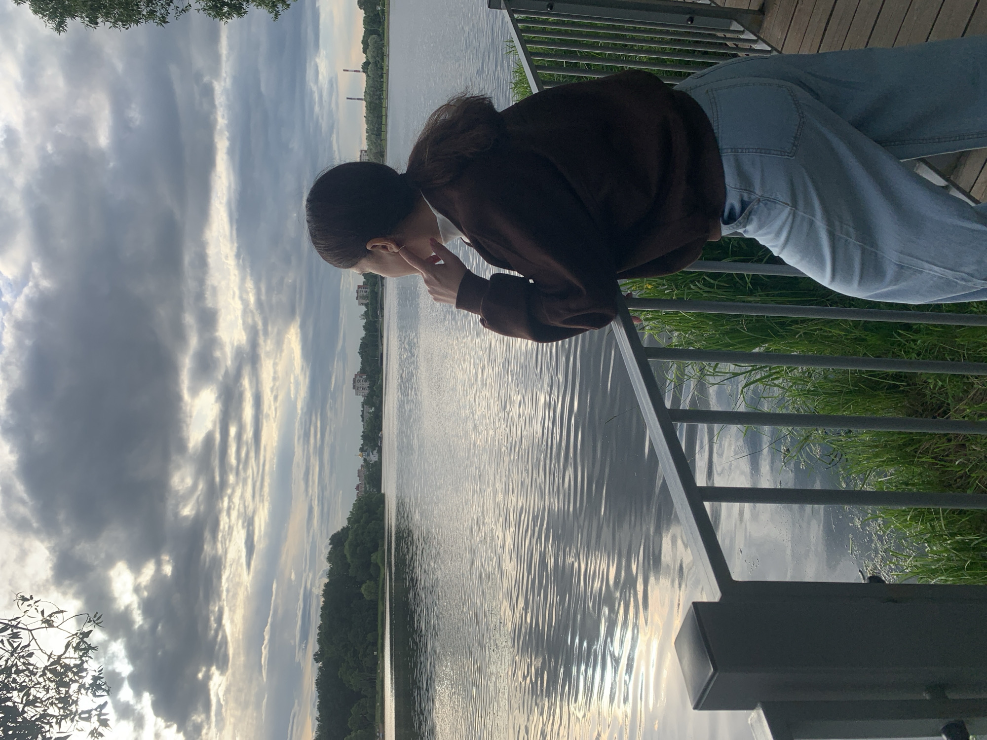 A person is leaning over a railing, looking out at a body of water. The person has dark hair pulled back and is wearing a brown hooded sweatshirt and light blue jeans. The person's face is obscured as they are looking down and slightly to the side, with one hand near their face. The scene appears to be outdoors, possibly a park or promenade, with a body of water in the background. The water has ripples, reflecting the sky, which is filled with dramatic clouds. There are trees and some buildings visible on the far bank of the water. The railing is made of dark metal, and there is wooden decking visible behind the person. The overall mood seems contemplative or perhaps curious. The lighting suggests it is late afternoon or early evening.