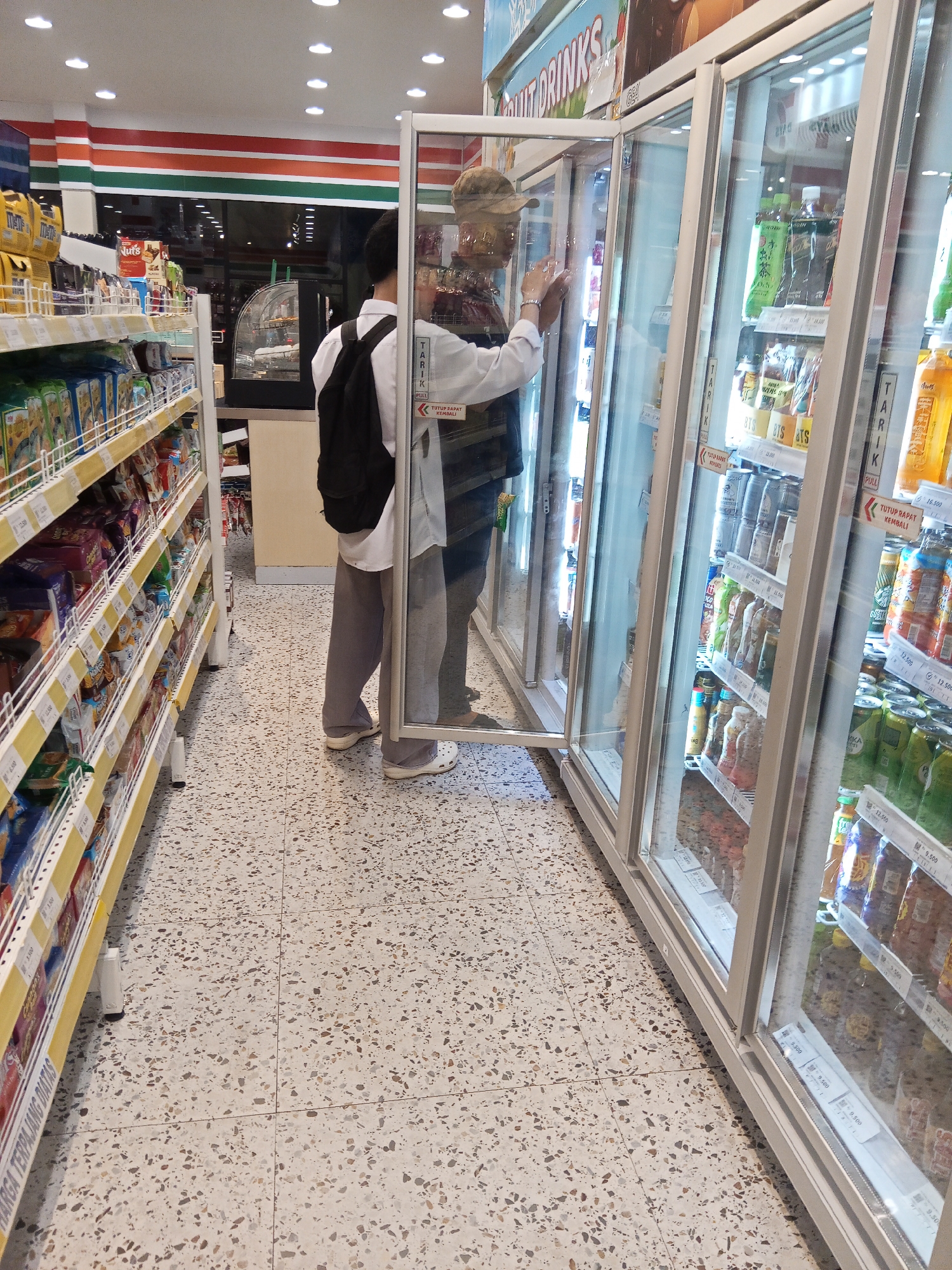 The image shows the interior of a Seven-Eleven convenience store, likely located in Lubuklinggau, Indonesia, given the context. Two individuals are present. One person, wearing a white shirt and grey pants, stands with their back to the camera, looking into a glass-door refrigerator display case. They are wearing a black backpack. Another person, wearing a cap and a patterned shirt, is partially visible on the right, also facing the refrigerator. The refrigerator is stocked with various bottled and canned beverages. To the left of the refrigerator are shelves filled with packaged snacks and other goods. The floor is tiled with a speckled pattern, and the ceiling is equipped with multiple circular lights. The store appears to be well-lit and organized.