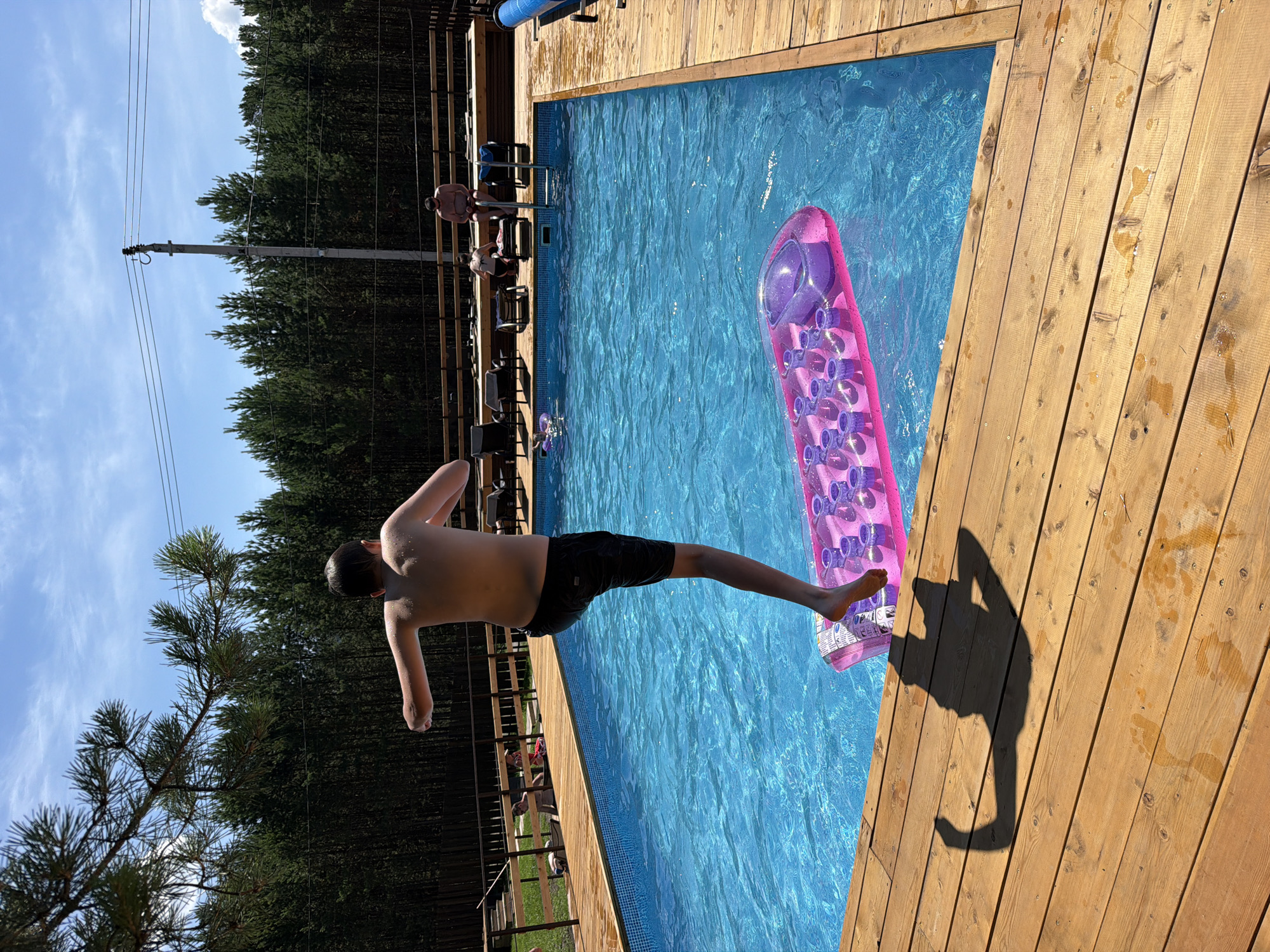 A young boy in black swim trunks is captured mid-air, jumping into a bright blue swimming pool from a wooden deck. His body is angled downwards, arms extended slightly to his sides, suggesting an imminent splash. A pink inflatable pool float, with numerous circular pockets, bobs on the water's surface nearby. The wooden deck, made of weathered planks, shows some water spots and foot imprints, indicating recent use.
In the background, other people are visible on the deck, some sitting on chairs and others standing. A wooden fence borders the pool area, beyond which a dense forest of dark green pine trees stretches towards a clear, bright blue sky with a few wispy clouds. The sunlight suggests it is daytime, likely midday or early afternoon, casting a strong shadow of the jumping boy onto the wooden deck. The overall scene conveys a sense of summer fun and outdoor recreation. There is no visible text in the image.
