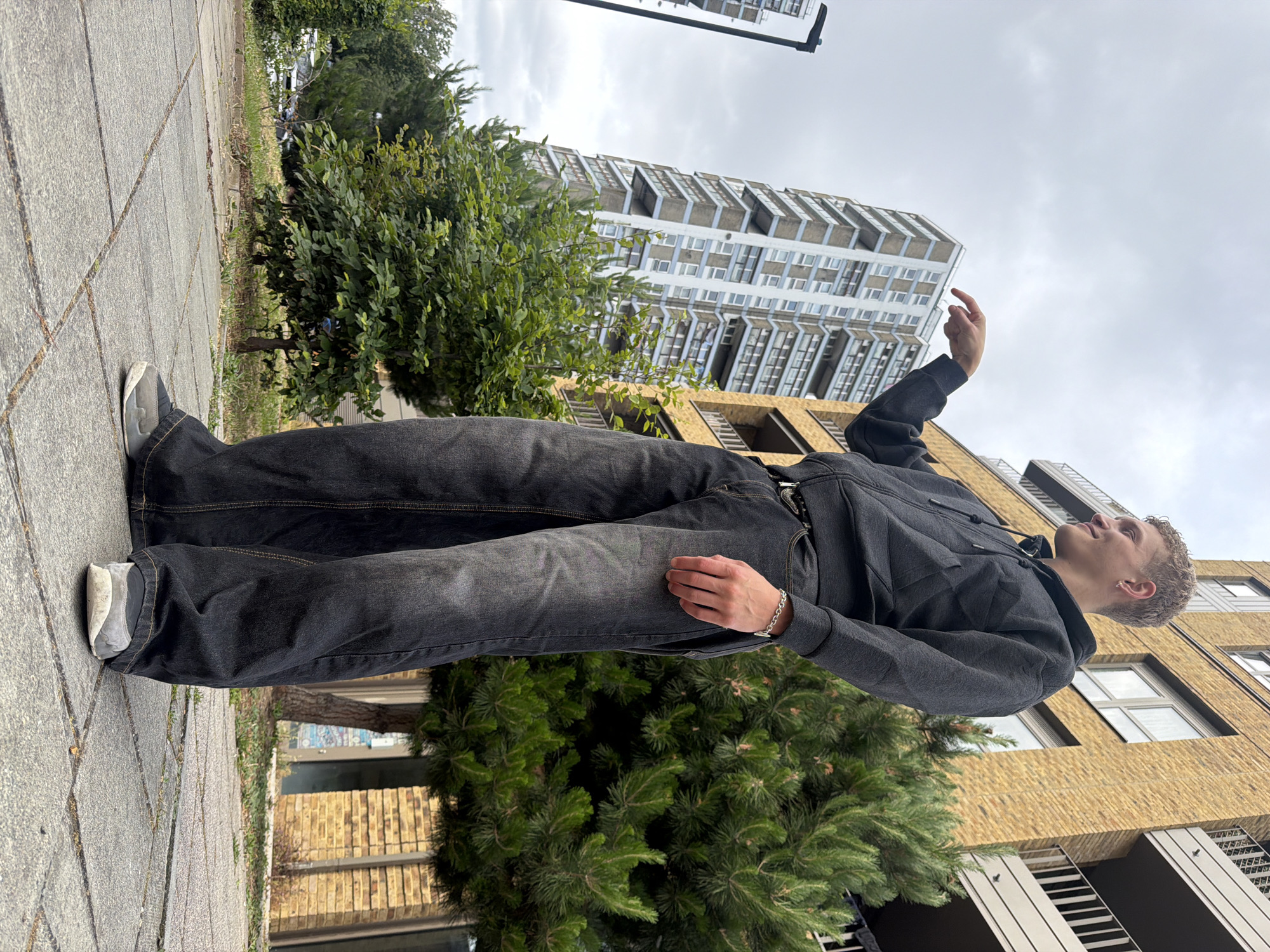 An outdoor, low-angle shot captures a young male in an urban environment on a cloudy day. The male, positioned center-right, has short blonde curly hair and fair skin, and is smiling while looking towards the left. He is wearing a dark grey zip-up hoodie, dark grey loose-fitting jeans, and grey and white sneakers. A silver chain bracelet is visible on his right wrist, and his left hand is extended, pointing towards the left side of the frame. He stands on a paved walkway consisting of square concrete slabs.
In the background to the left, a tall residential tower block rises, featuring a white facade with numerous balconies and what appear to be light blue or grey panels. To the right, partially obscured by the male, is a modern residential building constructed from yellow brick with dark-colored balconies and windows. Green foliage, including a prominent evergreen tree to the right of the male and smaller deciduous trees and bushes, fills the mid-ground. The specific architectural style of the buildings, particularly the tall tower and the yellow brick structure, is consistent with contemporary developments in the Elephant and Castle area of London, United Kingdom.