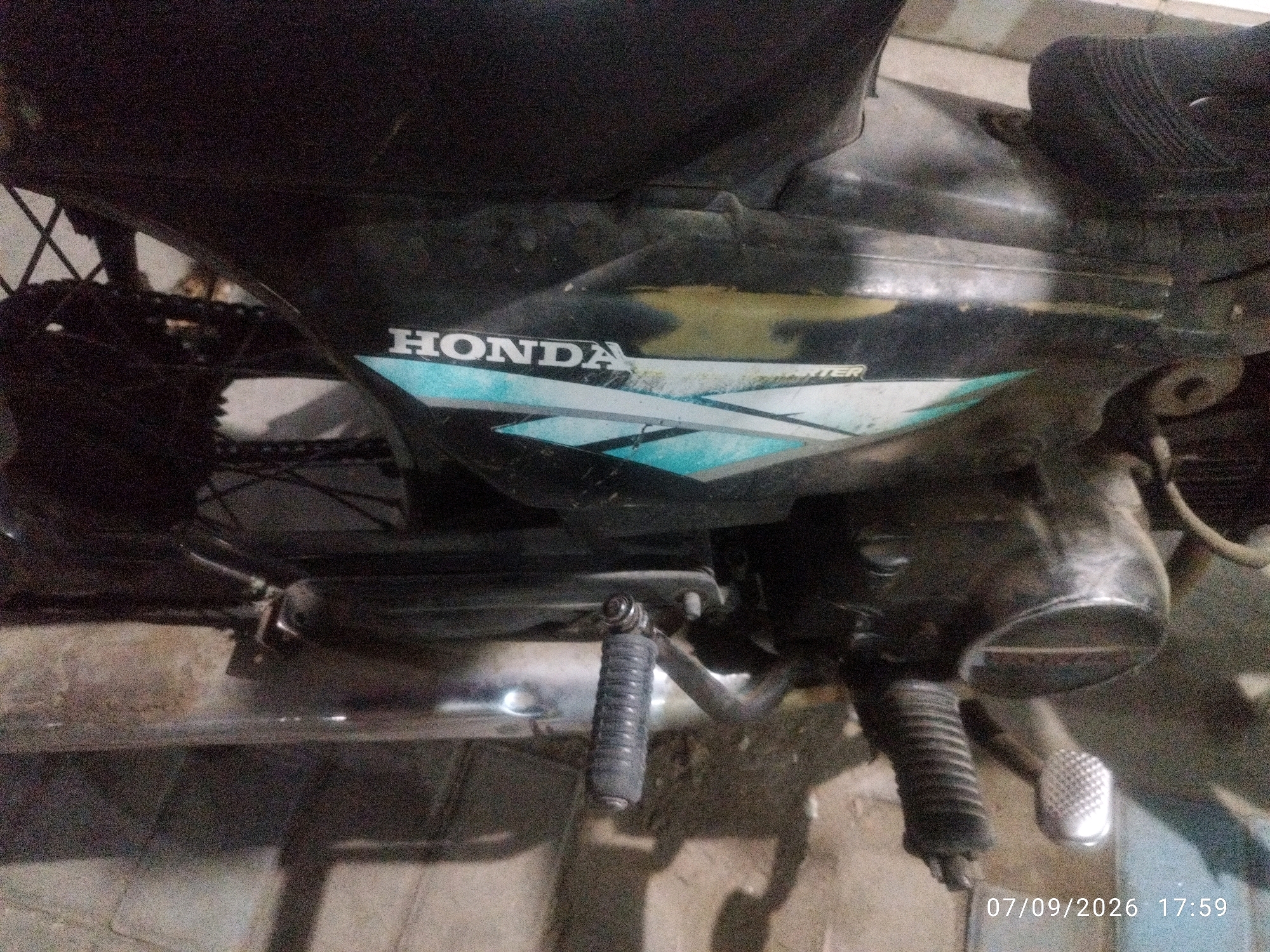 A close-up, overhead shot captures a section of a black Honda motorcycle, parked indoors on a tiled floor. The image focuses on the lower right side of the motorcycle, revealing the Honda logo and a portion of the bike's casing with blue and white graphics. Visible mechanical components include a kickstarter, a footpeg, and part of the chain and rear wheel assembly. The overall lighting is dim, suggesting an indoor or nighttime setting. The timestamp "07/09/2026 17:59" is overlaid on the bottom right of the image. The context of Prajuritkulon, Indonesia, is not discernible from the visual information present in the image.