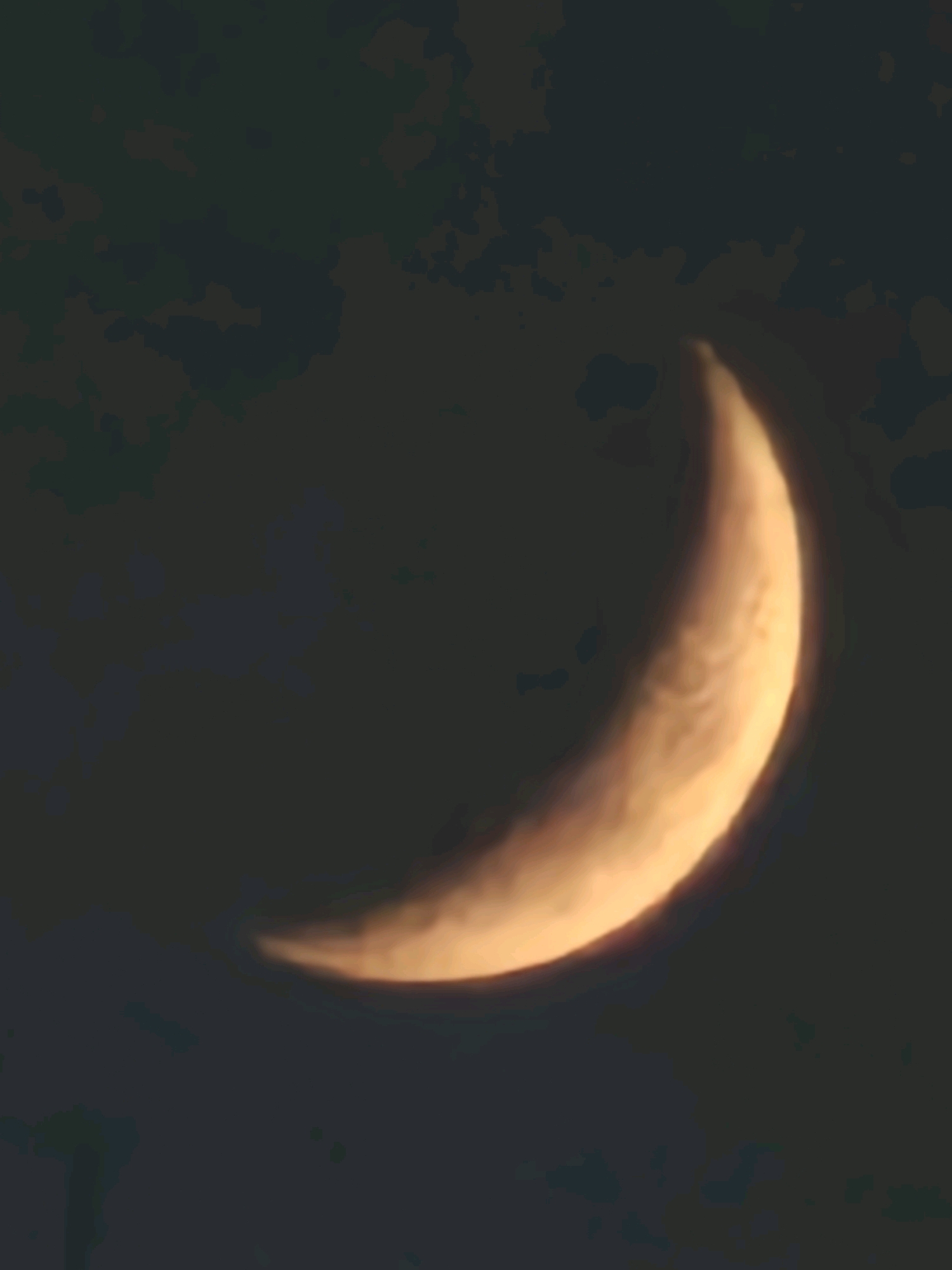 The image shows a crescent moon against a dark, cloudy sky. The moon is illuminated from the right, with its left side in shadow. The surface of the moon shows some detail, including what appear to be craters or variations in texture. The clouds are dark and indistinct, creating a mottled background. The image is a photograph taken at night. There are no people, objects, or any indication of the city of Al Kuwayt, Kuwait in the image.