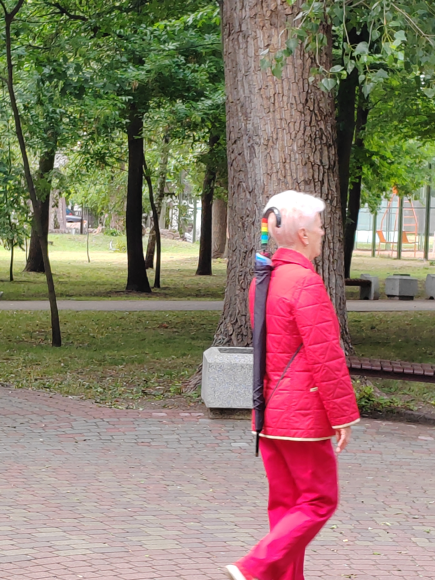 An elderly woman with short, white hair, wearing a red quilted jacket and red pants, walks along a paved path in a park. An umbrella, attached to a black strap over her shoulder, hangs down her back. The umbrella has a rainbow-colored handle. She appears to be walking to the right of the frame. The park features mature trees, grass, and benches. In the background, a playground structure is visible. The scene is consistent with a public park area within the city of Brest, Belarus.