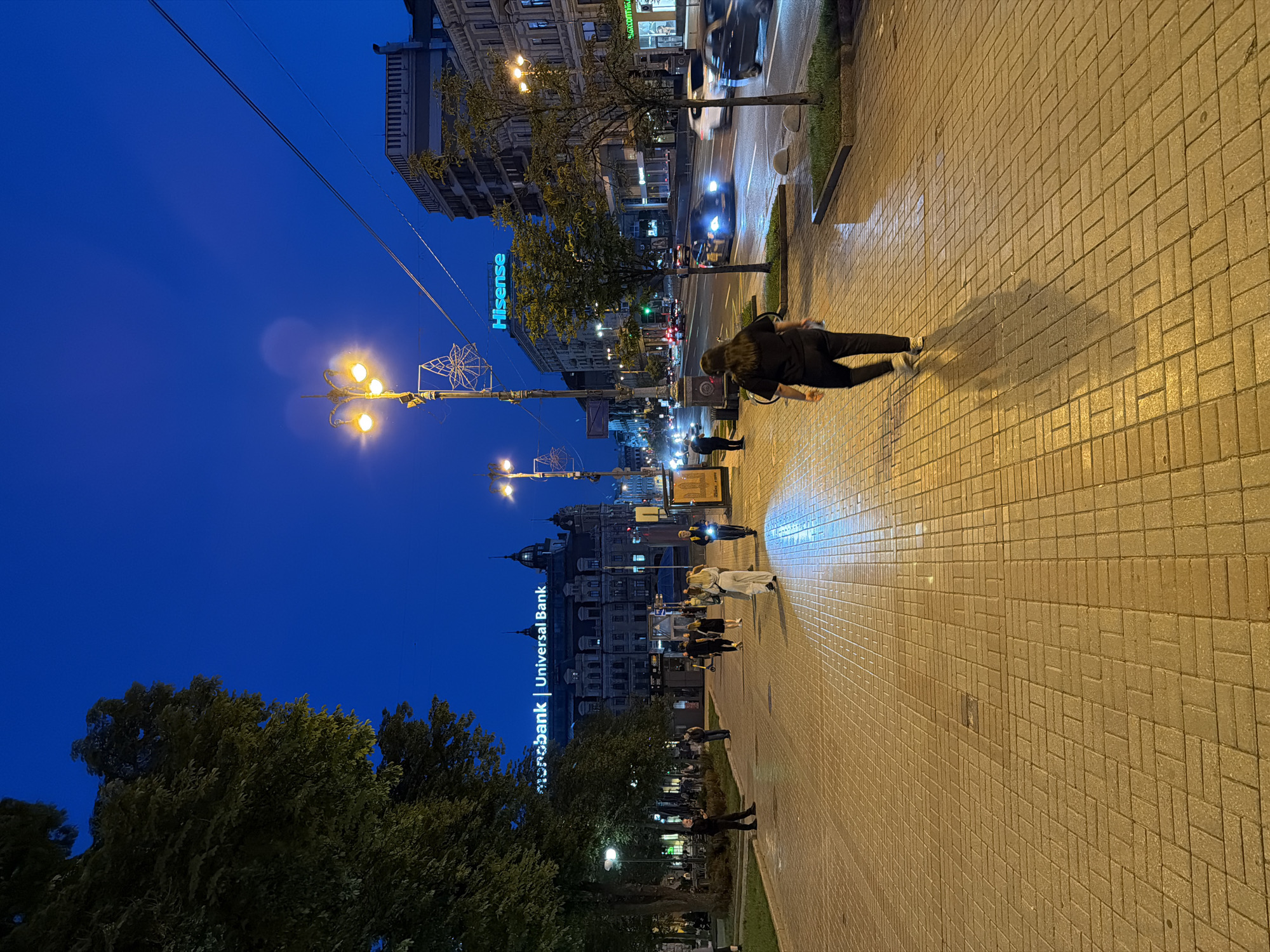 The image depicts an evening scene on a wide, paved pedestrian walkway in Kyiv, Ukraine, likely on Khreshchatyk Street or a similar central avenue, given the prominent advertising and urban architecture. The sky is a deep twilight blue, indicating the transition from day to night.
On the left side of the walkway, large trees with dense foliage cast shadows, and several people are visible sitting at outdoor cafe tables. Further down the walkway, more pedestrians are walking, some in small groups. One individual in the foreground is walking away from the camera, casting a long shadow on the light-colored, patterned paving stones. The paving stones appear slightly wet, reflecting the ambient light from streetlamps and buildings.
The right side of the walkway borders a busy street with moving vehicles, their headlights and taillights creating streaks of light due to motion blur. Several buildings line the street, some featuring illuminated signage. Notably, a large building in the background displays the "monobank | Universal Bank" logo in glowing neon letters, and another building has a prominent "Hisense" sign. Streetlights with a warm yellow glow are strategically placed along the walkway, illuminating the path. The overall atmosphere is one of an active urban environment at dusk.