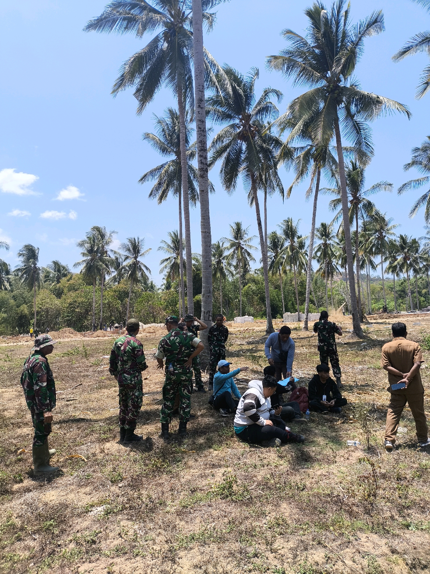 In Modelomo, Indonesia, a group of individuals, predominantly in military camouflage uniforms, are gathered in an open, dry, grassy field dotted with scattered palm trees. The scene appears to be a construction or development site, with visible piles of building materials, including stacks of white blocks and rebar protruding from the ground in the background. Several individuals are seated on the ground, engaged in conversation or looking at their phones. Two individuals in military uniforms are standing and observing the situation, while others are in the process of walking or standing. The environment is characterized by a clear blue sky with a few scattered white clouds, and a dense backdrop of lush green trees, primarily palm trees, suggesting a tropical climate. The ground is dry and appears to be covered in a mixture of dry grass, dirt, and small plants. A man in a brown uniform is standing to the right, holding a phone. The overall impression is one of a casual gathering or a briefing session amidst a partially cleared or developing area.