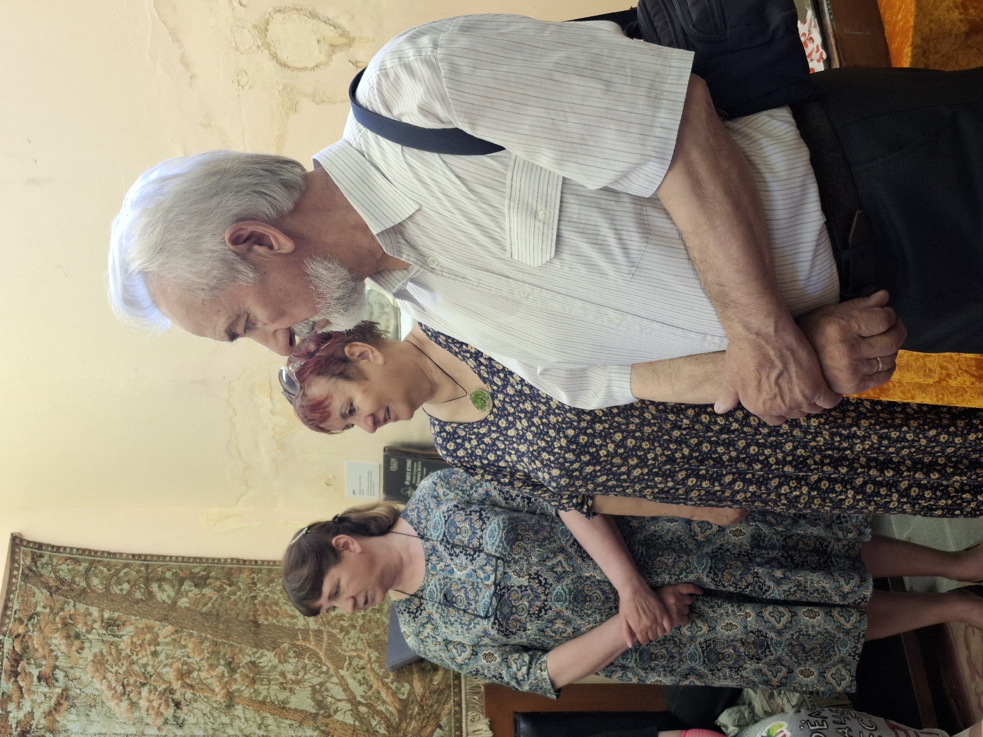 Three individuals are indoors in a room with a tapestry on the wall. The location is Марковский, Russia. An older man with gray hair and a white striped shirt stands at the top of the composition. Below him is a woman with red hair wearing a dark floral dress. At the bottom, another woman in a blue patterned dress stands with her hands clasped in front of her. All three appear to be looking down.