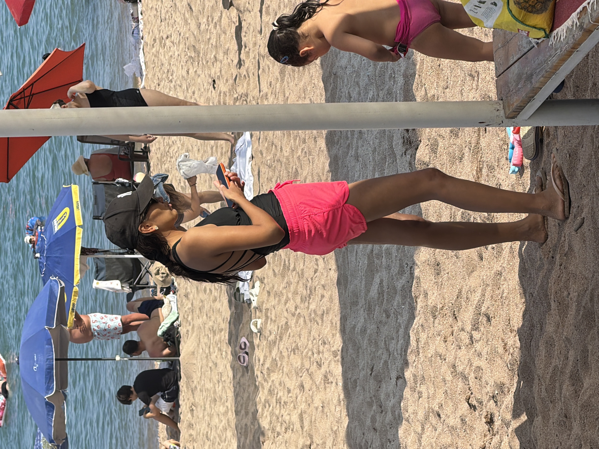 In the city of Unknown, Unknown, on a sandy beach, a young woman in a black swimsuit and bright pink shorts stands and looks at her phone. She wears a black baseball cap. To her right, a young child in a pink swimsuit plays, partially obscured by a pole. Other beachgoers are visible in the background, some under blue and orange umbrellas. The water of a lake or ocean is behind them. Scattered on the sand are various flip-flops and a bench.