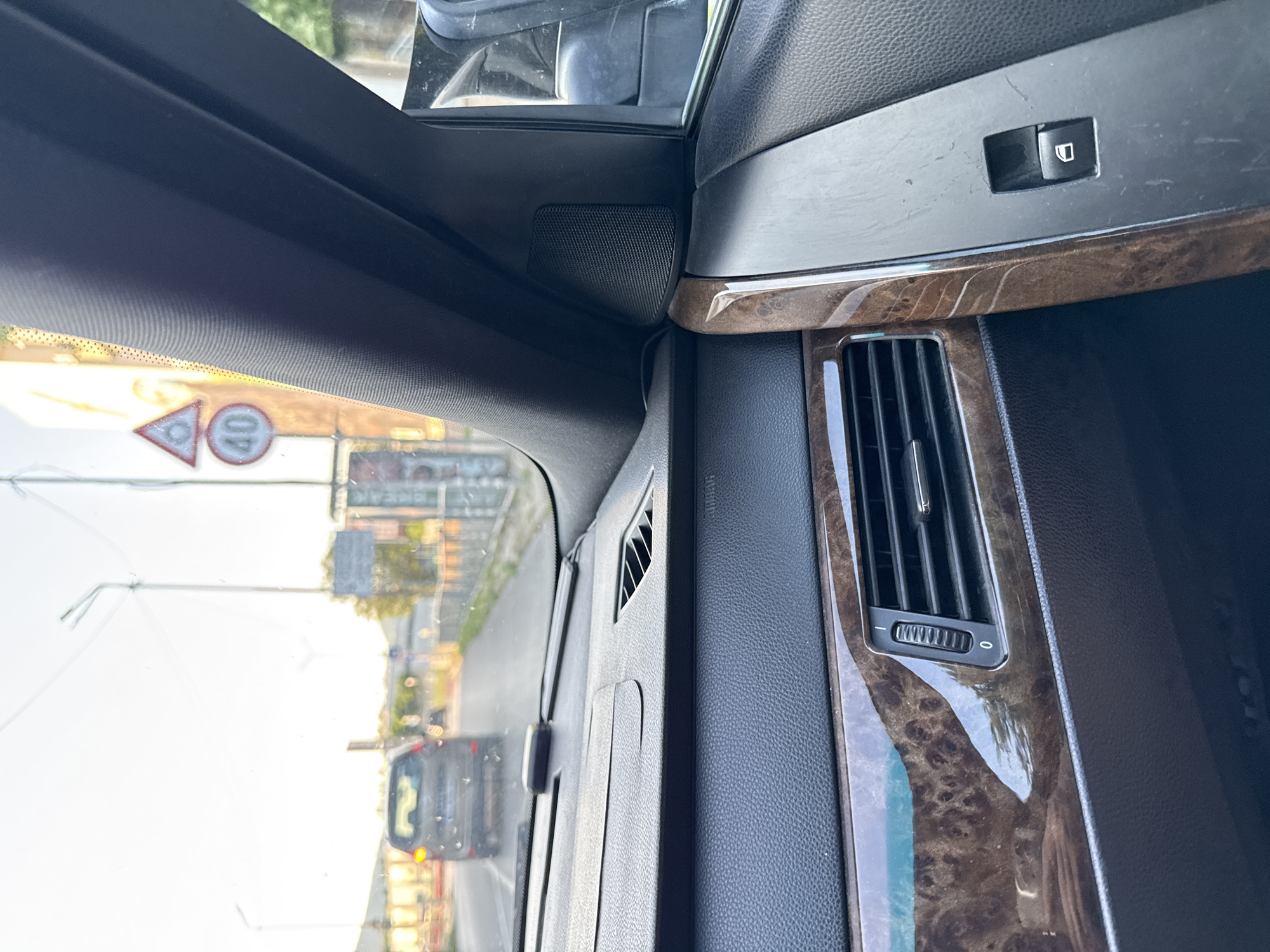 The image depicts the interior of a vehicle, specifically from the front passenger's perspective, looking forward through the windshield. The scene is located in Stara Zagora, Bulgaria.
The car's dashboard, visible in the foreground, features dark textured plastic surfaces and prominent burlwood-style trim along the lower dash and door panel. A central air vent with a vertical adjuster for airflow direction and a horizontal dial (marked "0" to "I") for intensity is visible on the passenger side. Above the lower dashboard, a smaller air vent is present on the upper part. A small, dark electronic device is mounted on the dashboard near the windshield. The A-pillar is covered in dark fabric, and a speaker grille is integrated into the panel near the side mirror. A window control switch is visible on the passenger door panel.
Beyond the windshield, a street scene unfolds under bright, hazy daylight, possibly late afternoon or early morning, given the soft, warm light. An asphalt road, possibly slightly wet or reflective, extends forward. A light-colored sedan is visible ahead, traveling in the same direction. On the right side of the road, a series of buildings and streetlights line the thoroughfare. Two traffic signs are prominent on the right: a red-bordered triangular warning sign (contents unclear due to blur/angle) positioned above a circular regulatory sign indicating a "40" speed limit (kilometers per hour). No individuals are distinctly visible within the frame.