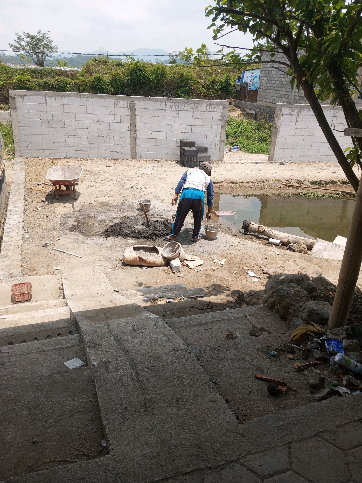 In the city of Kalibeber, Indonesia, a construction site is depicted under a bright, overcast sky. In the foreground, concrete stairs lead up to a work area where a man is actively engaged in mixing cement. He is wearing a white long-sleeved shirt with blue trim and dark pants, and is standing near a pile of mixed cement, using a shovel. Nearby, a bag of cement is partially emptied, and several buckets and mixing tools are scattered on the sandy ground. A red wheelbarrow sits to the left, with a basket nearby. Behind the man, a body of water is visible, with a fallen log and a white pipe at its edge. Construction materials, including white cinder blocks, are stacked and form partial walls around the site. Lush green vegetation lines the background, with hills and buildings visible in the distance. The scene suggests ongoing construction work in a semi-rural environment.